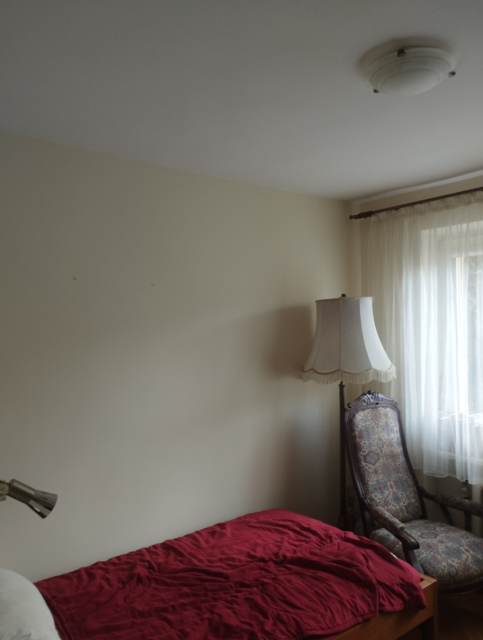 Pokój po odświeżeniu: kremowe ściany, czerwona narzuta na łóżku, ozdobny fotel i lampa podłogowa. Widoczne ślady po niedawnych pracach.