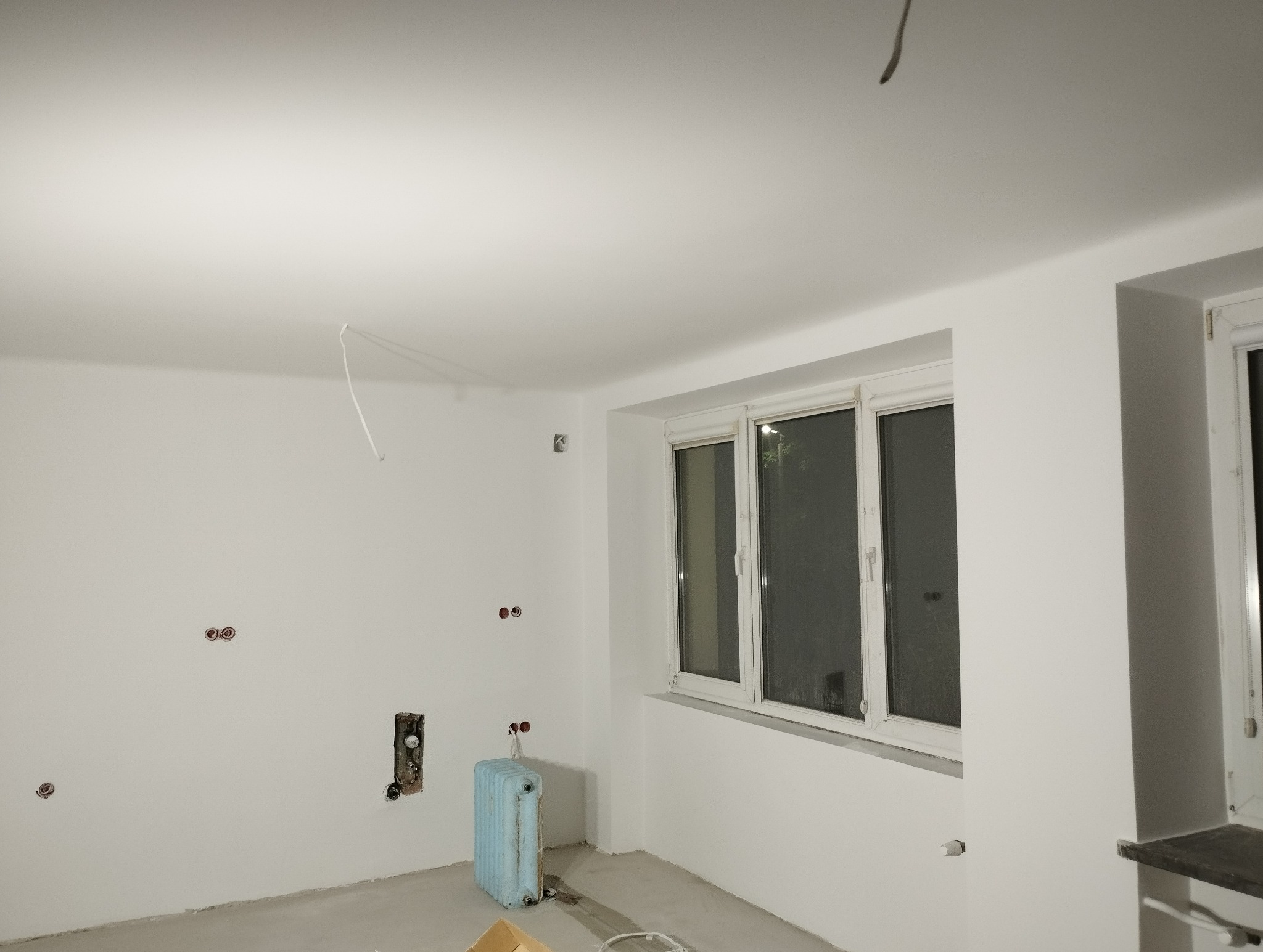 Białe ściany po malowaniu w pokoju z oknem i grzejnikiem. Widoczne gniazdka elektryczne i przewody. Świeżo pomalowane wnętrze w trakcie wykańczania.