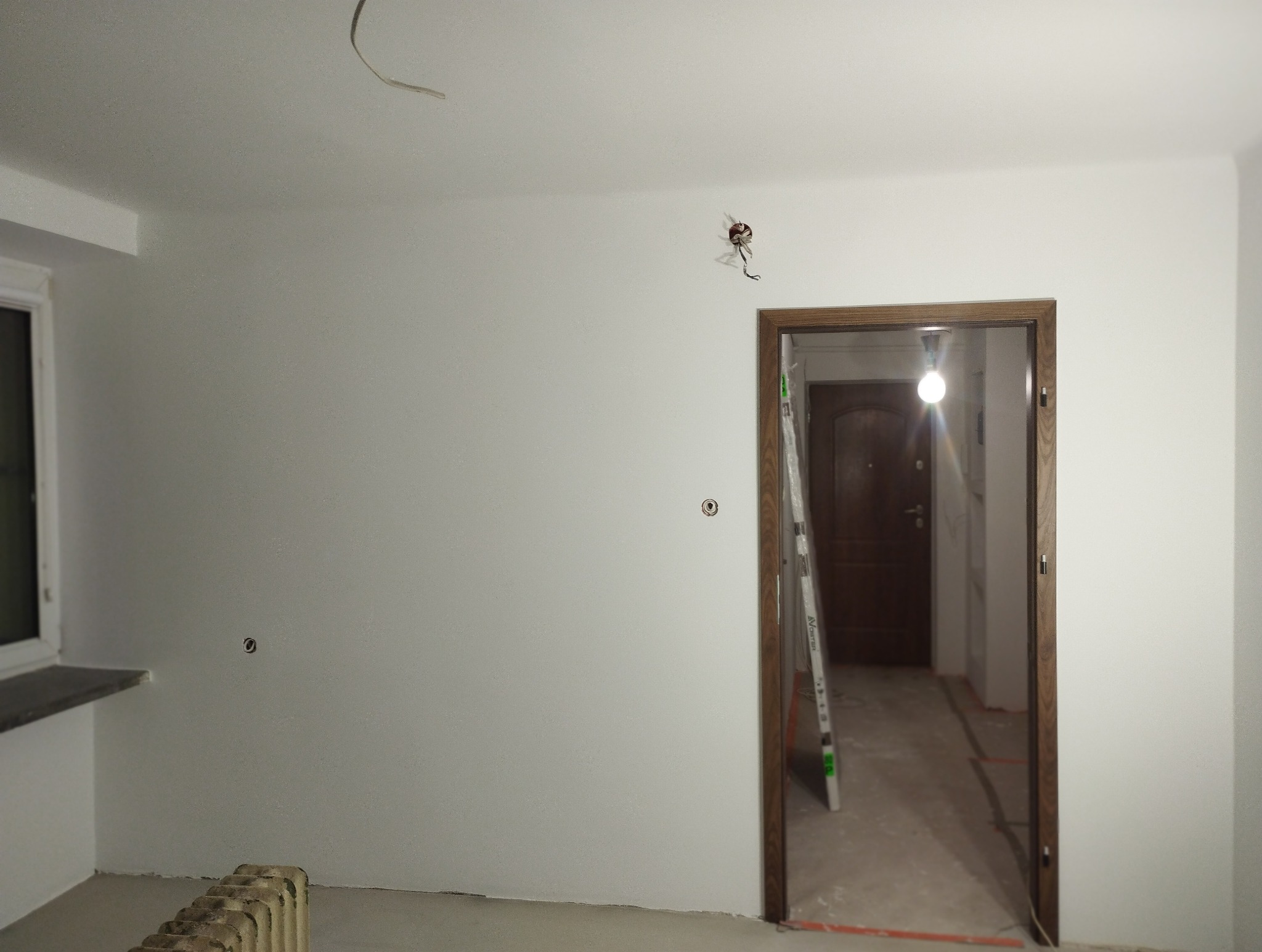 Wnętrze po odświeżeniu: białe ściany, widoczne gniazdka, drabina oparta o futrynę drzwi. Grzejnik pod oknem. Minimalistyczny wystrój, świeżo po pracach wykończeniowych.