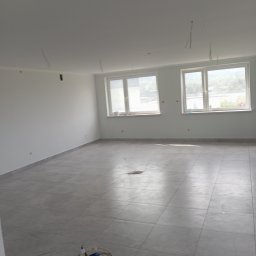 HK WYKOŃCZENIA WNĘTRZ KAMIL HAŃDEREK - Pusty, jasny pokój z szarymi płytkami podłogowymi, świeżo pomalowanymi ścianami z wystającymi przewodami elektrycznymi z sufitu, widoczne dwa okna z widokiem na zewnątrz i przedłużacz leżący...