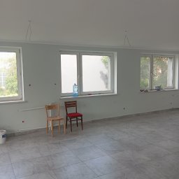HK WYKOŃCZENIA WNĘTRZ KAMIL HAŃDEREK - Pomalowane na jasno pomieszczenie z trzema oknami, szarą podłogą z płytek, dwoma krzesłami pod oknem, widocznymi przewodami elektrycznymi wystającymi z sufitu i śladami po szpachlowaniu na ścianach.