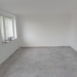HK WYKOŃCZENIA WNĘTRZ KAMIL HAŃDEREK - Pusty, jasny pokój z szarymi płytkami podłogowymi imitującymi marmur, świeżo pomalowane ściany na biało, widoczne rury grzewcze i puszka z farbą na parapecie okna.