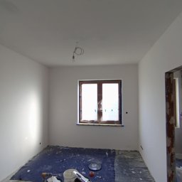 HK WYKOŃCZENIA WNĘTRZ KAMIL HAŃDEREK - Pokój w trakcie prac wykończeniowych: świeżo pomalowane białe ściany i sufit, zabezpieczona folią podłoga, widoczne wiaderka z farbą i narzędzia malarskie, okno zabezpieczone folią malarską.