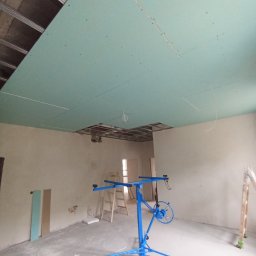 HK WYKOŃCZENIA WNĘTRZ KAMIL HAŃDEREK - Montaż zielonej płyty karton gips na suficie z użyciem podnośnika. Widoczne profile sufitowe i fragmenty niezabudowane. W tle drabina i fragmenty płyt.