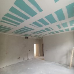 HK WYKOŃCZENIA WNĘTRZ KAMIL HAŃDEREK - Wnętrze w trakcie prac: sufit z płyt kartonowo-gipsowych, częściowo zaszpachlowany na niebiesko, ściany przygotowane do dalszych prac wykończeniowych, widoczna drabina.