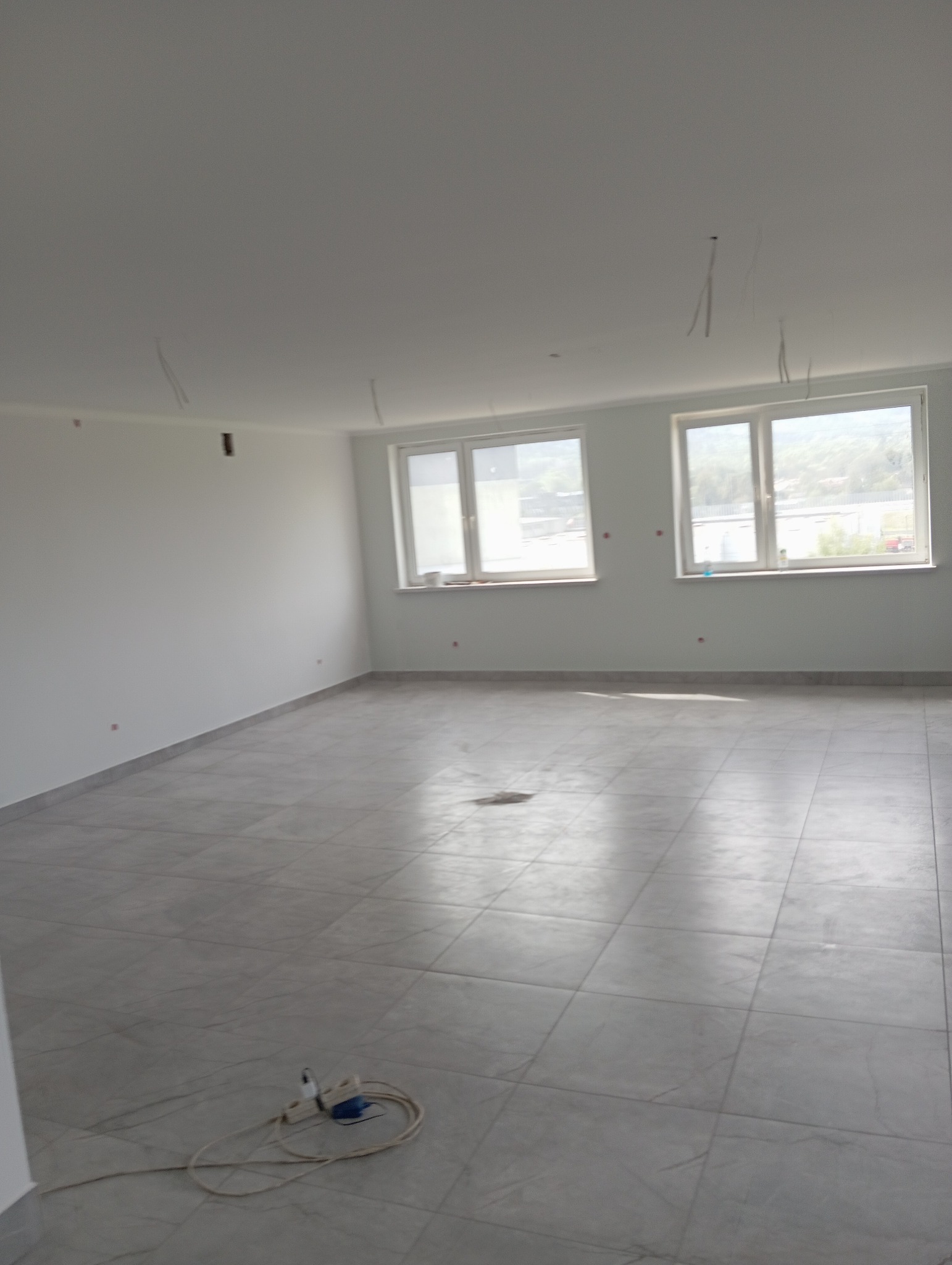 Pusty, jasny pokój z szarymi płytkami podłogowymi, świeżo pomalowanymi ścianami z wystającymi przewodami elektrycznymi z sufitu, widoczne dwa okna z widokiem na zewnątrz i przedłużacz leżący...
