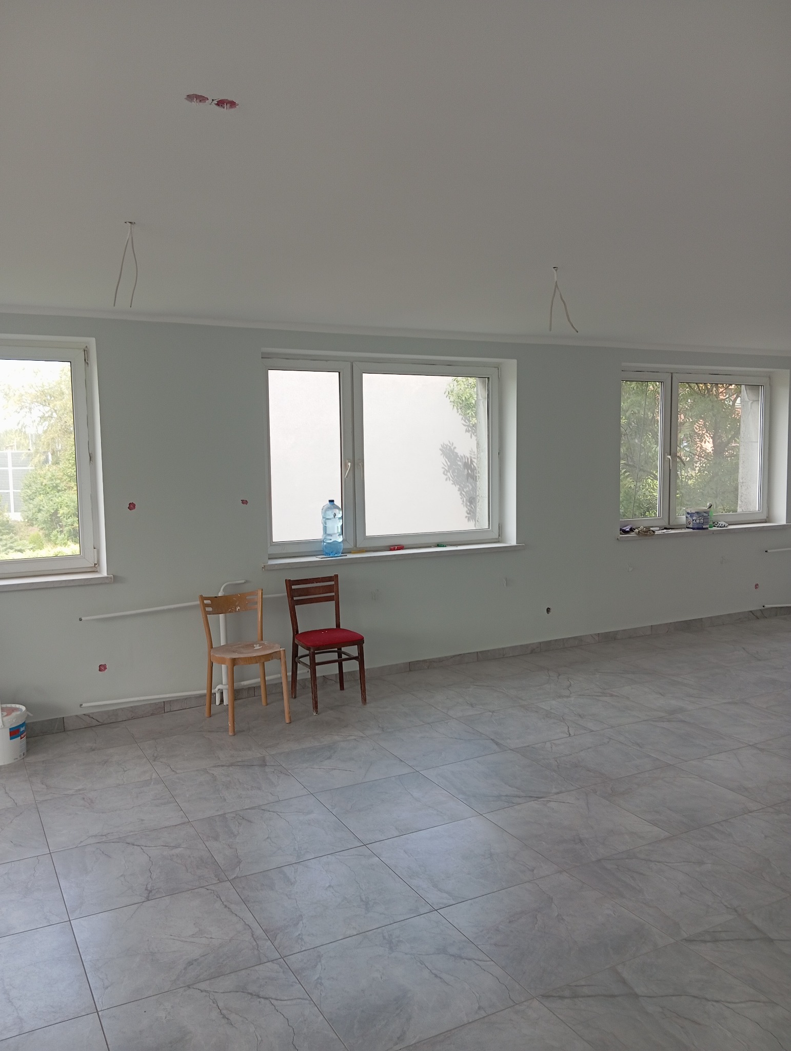 Pomalowane na jasno pomieszczenie z trzema oknami, szarą podłogą z płytek, dwoma krzesłami pod oknem, widocznymi przewodami elektrycznymi wystającymi z sufitu i śladami po szpachlowaniu na ścianach.