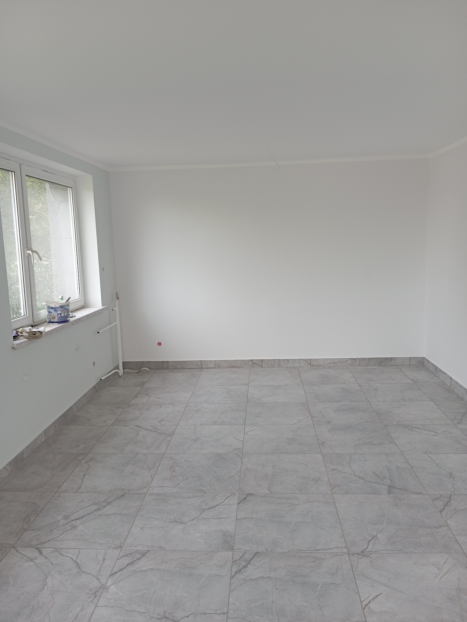 Pusty, jasny pokój z szarymi płytkami podłogowymi imitującymi marmur, świeżo pomalowane ściany na biało, widoczne rury grzewcze i puszka z farbą na parapecie okna.