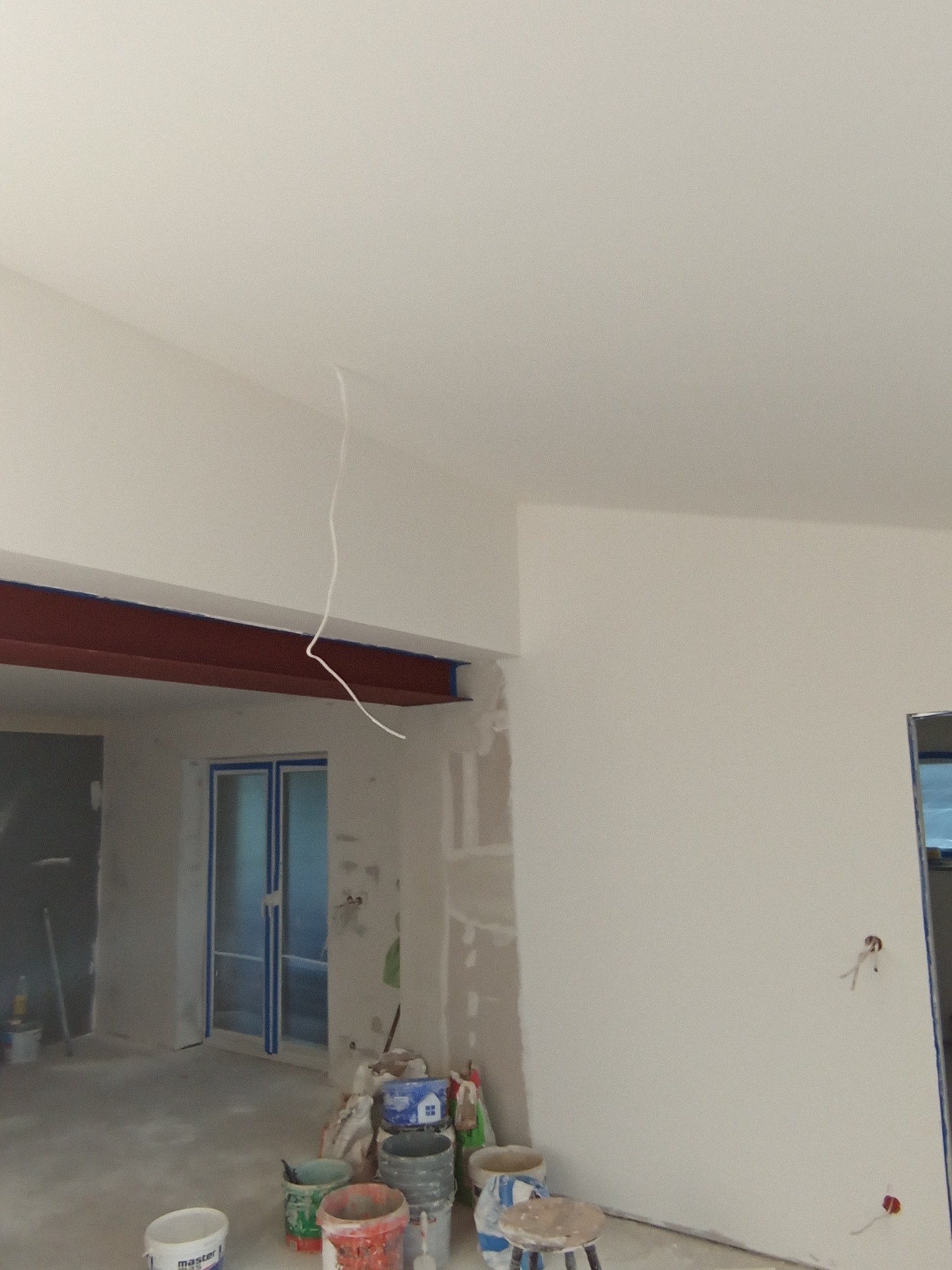 Wnętrze w trakcie prac przygotowawczych do wykończenia: ściany po nałożeniu gładzi, zabezpieczone okna i framugi taśmą malarską, widoczne wiadra z zaprawą i narzędzia.