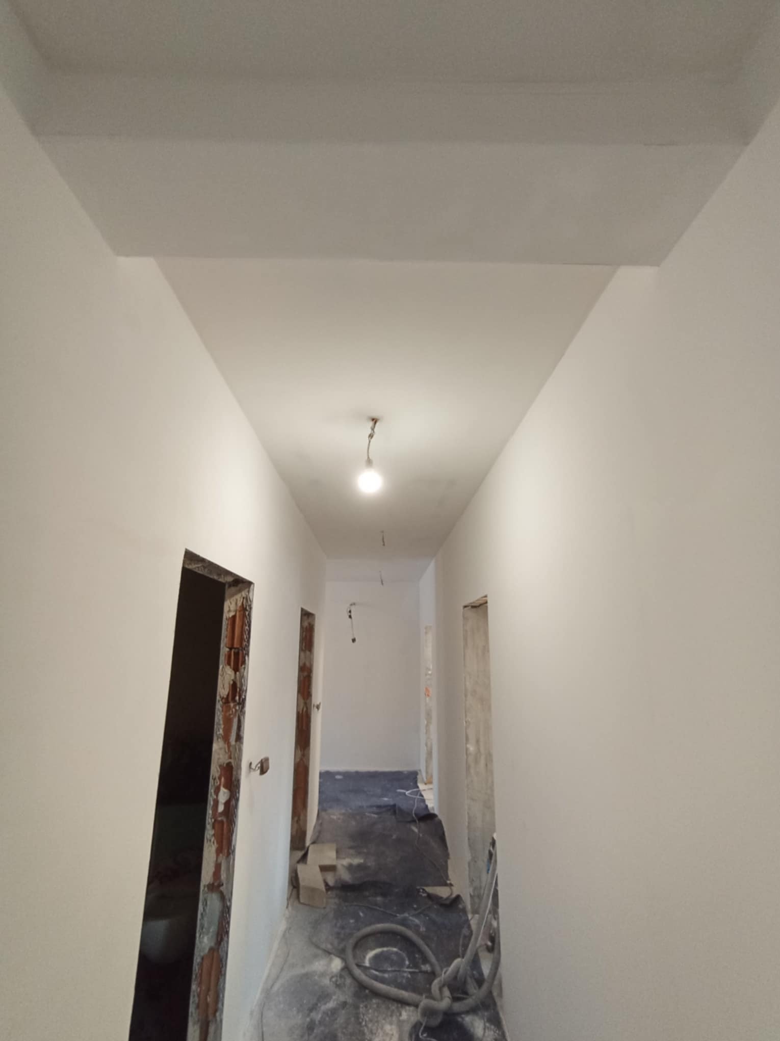Korytarz w trakcie prac wykończeniowych, ściany pokryte świeżo nałożoną gładzią, widoczne ościeżnice drzwiowe w stanie surowym oraz częściowo zakryta podłoga.