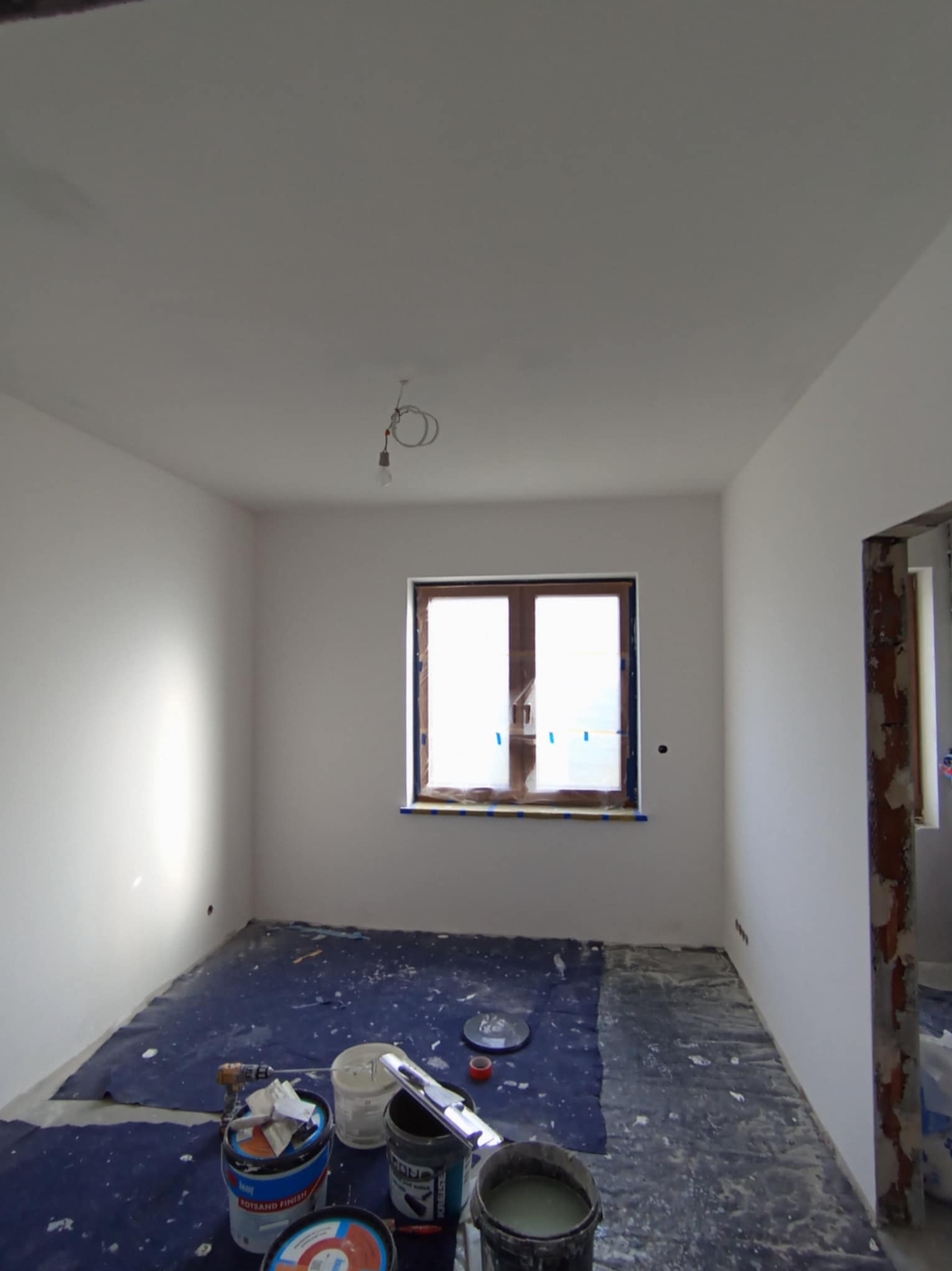 Pokój w trakcie prac wykończeniowych: świeżo pomalowane białe ściany i sufit, zabezpieczona folią podłoga, widoczne wiaderka z farbą i narzędzia malarskie, okno zabezpieczone folią malarską.
