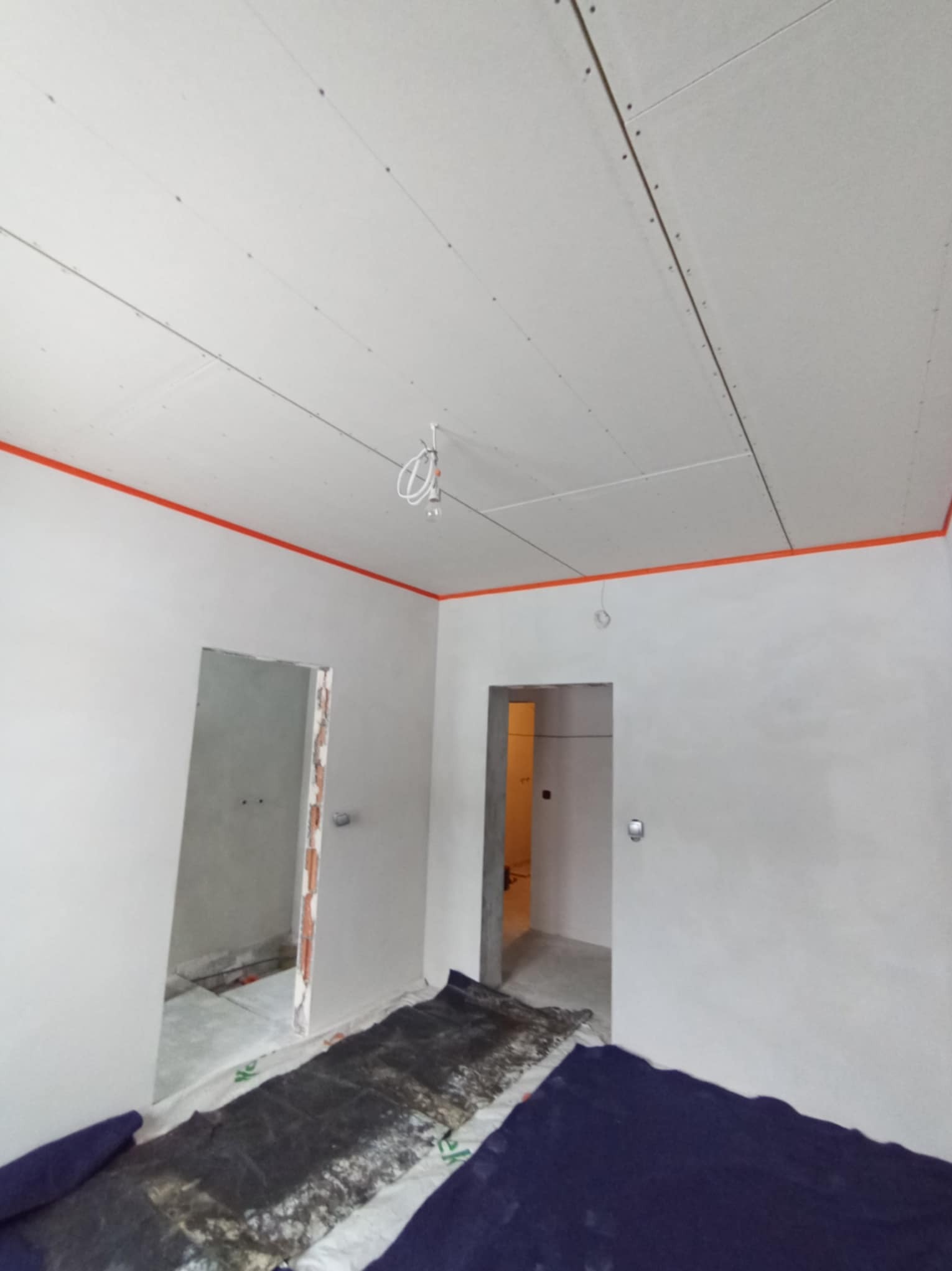 Pokój w trakcie prac: zamontowane płyty gipsowo-kartonowe na suficie, zabezpieczone folią podłogi, widoczne kable oświetleniowe.