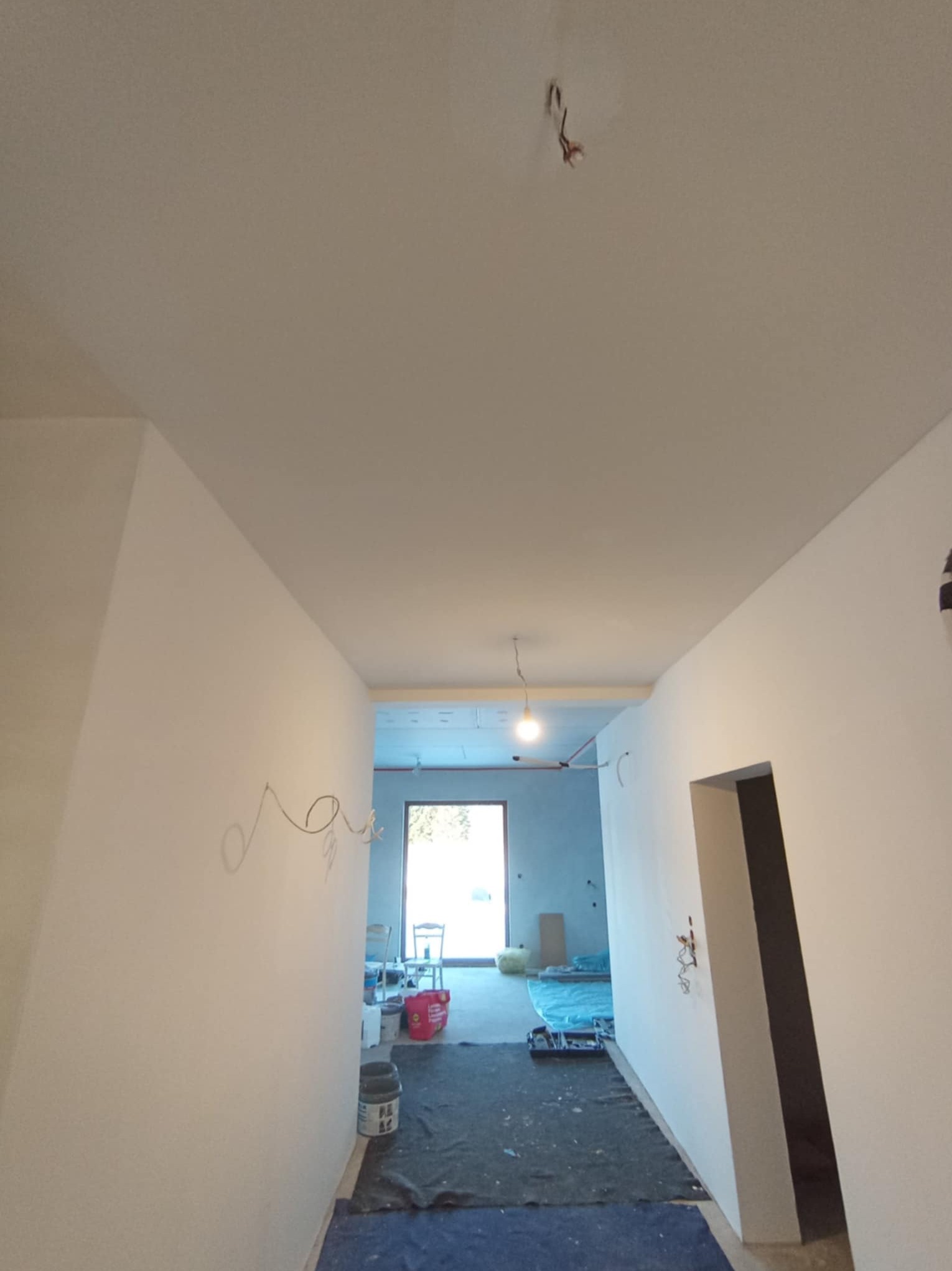 Korytarz w trakcie malowania ścian na biało, widoczne niedokończone prace, kable wystające ze ściany i sufitu, zakryta podłoga, wiadro z farbą przy ścianie.