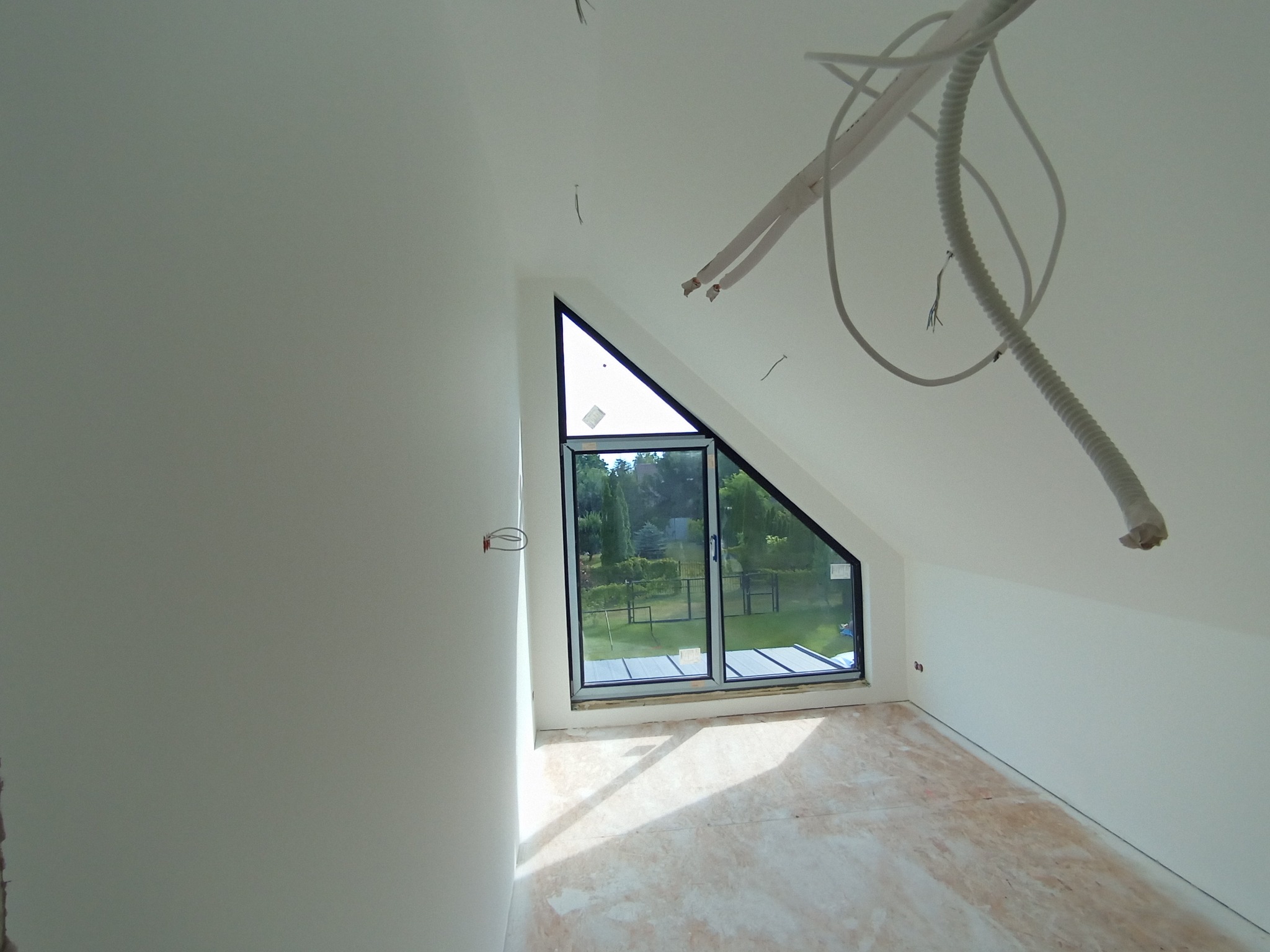 Jasne wnętrze poddasza z trójkątnym oknem, świeżo pomalowane na biało, z widocznymi przewodami na suficie i przygotowanym podłożem pod podłogę.