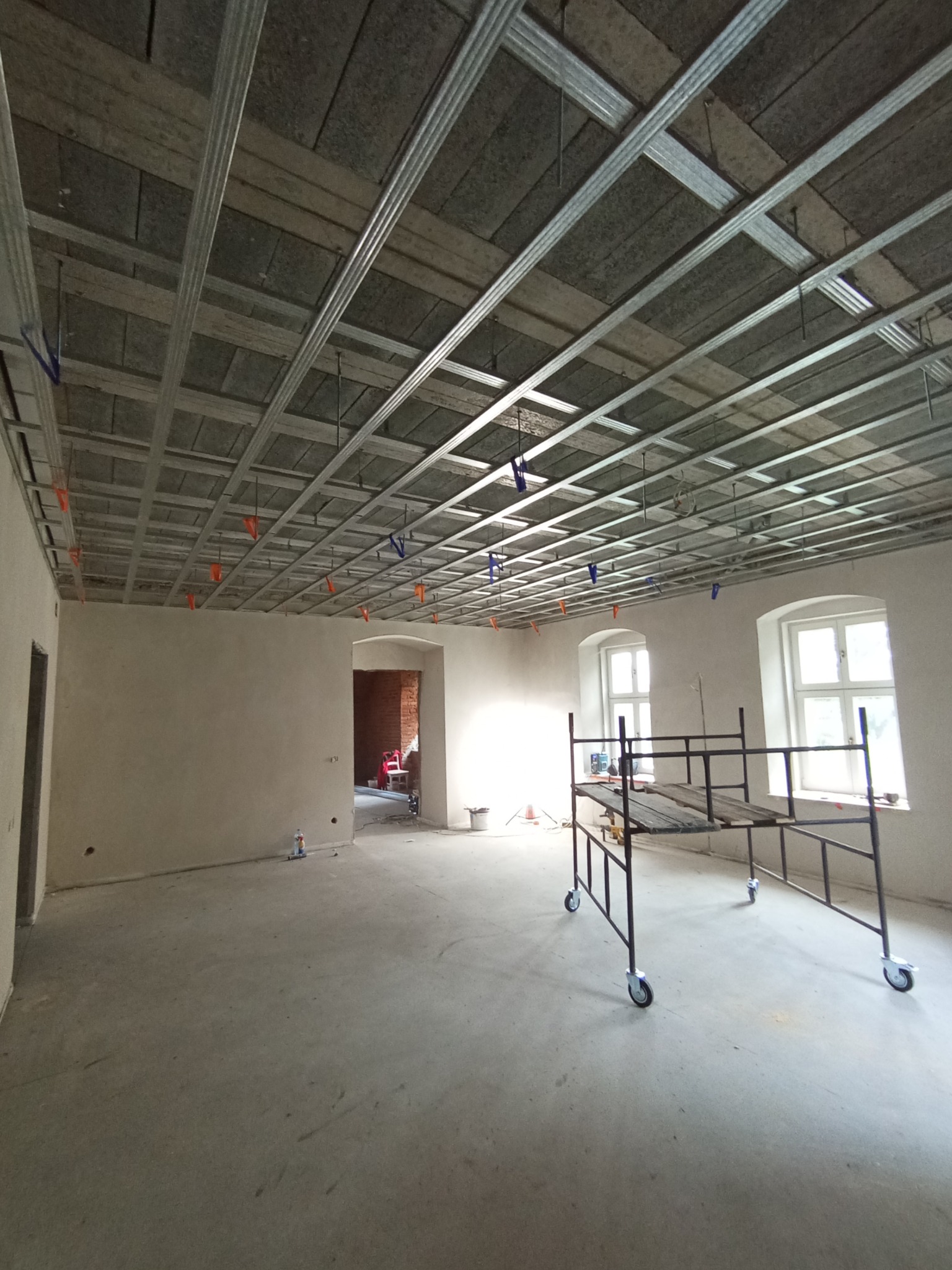 Montaż stelaża sufitu podwieszanego z płyt karton-gips w przestronnym wnętrzu. Widoczne rusztowanie i przygotowane ściany. Charakterystyczne okna z łukami.