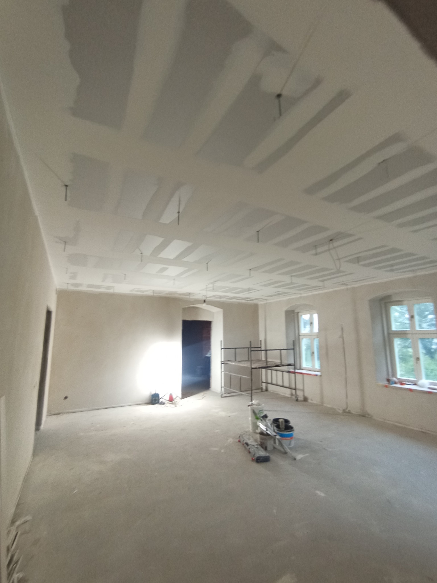 Pokój w trakcie prac wykończeniowych: sufit z płyt karton-gips, ściany po szpachlowaniu, widoczne narzędzia i rusztowanie. Dwa okna z widokiem na zieleń.