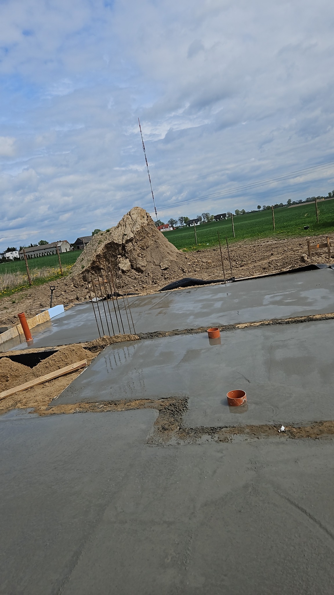 Wylany fundament z wystającymi prętami zbrojeniowymi na tle sterty ziemi i zielonego pola w słoneczny dzień.