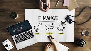 Ręka pisząca na plakacie z napisem FINANCE i rysunkami przedstawiającymi proces finansowy: inwestycje, oszczędności i zyski. Obok laptop, aparat fotograficzny, filiżanka kawy i kartki na drewnianym...