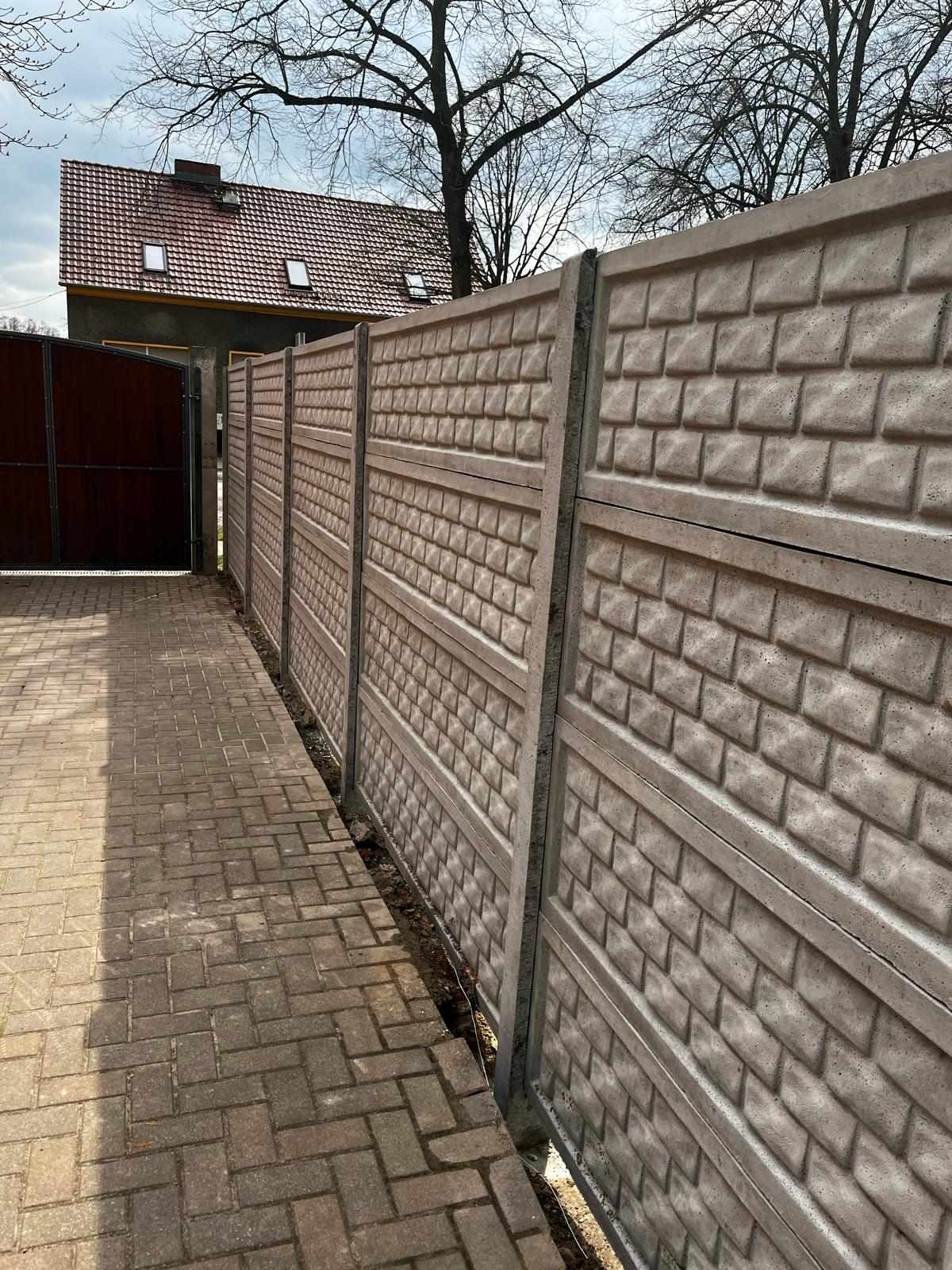 Nowo postawiony, szary, betonowy płot z imitacją cegieł, widok z boku, z kostki brukowej ułożony chodnik prowadzący do drewnianej bramy.