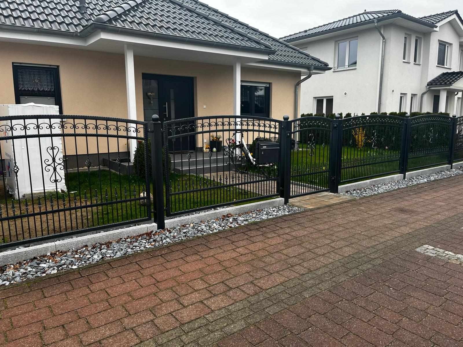 Czarna, metalowa brama i ogrodzenie posesji z dekoracyjnymi elementami, widoczne z poziomu brukowanego chodnika, z domem w tle.