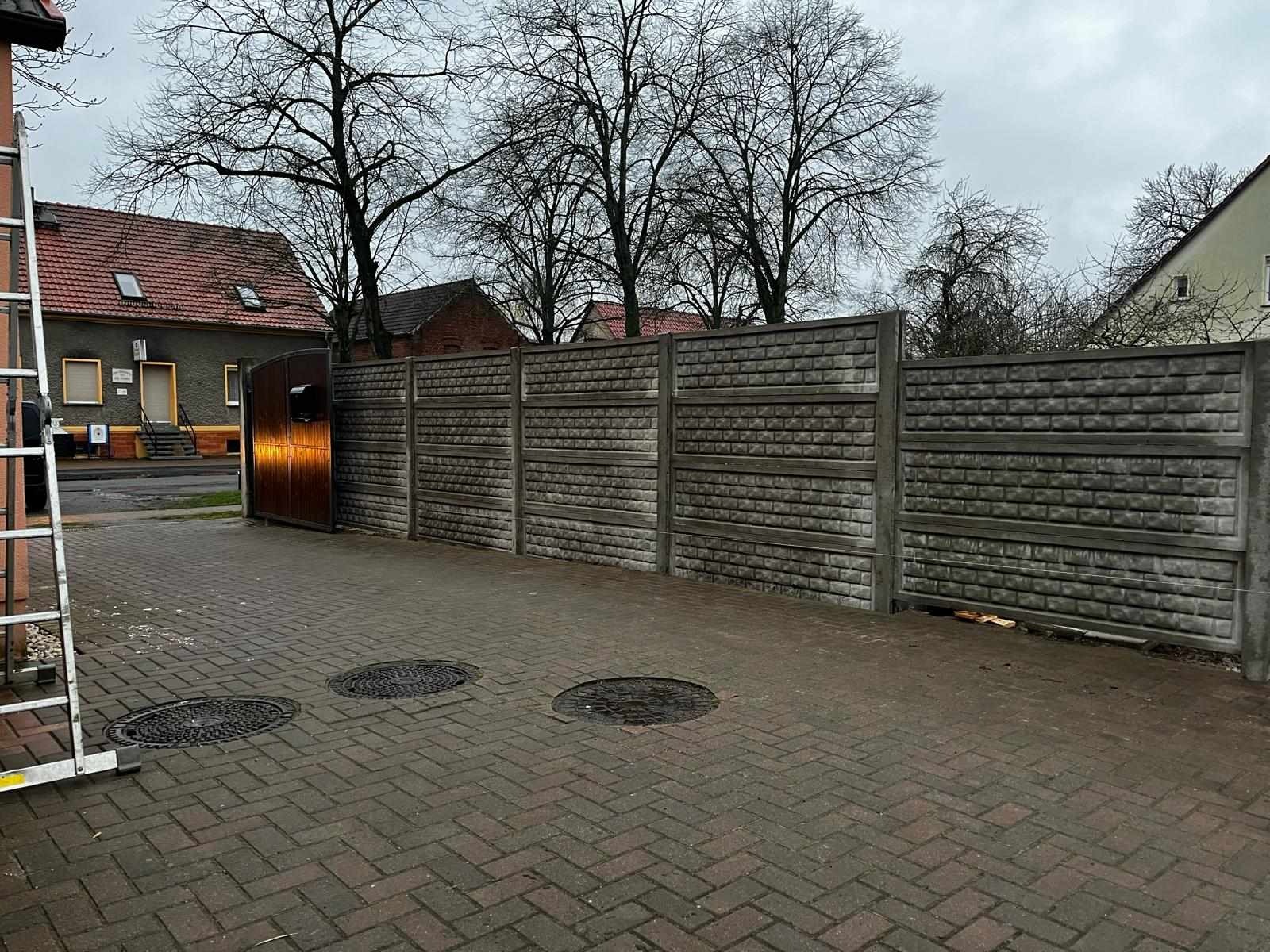 Ułożona kostka brukowa na podjeździe z trzema studzienkami kanalizacyjnymi, widoczny betonowy płot panelowy i metalowa brama w tle, pochmurne niebo.