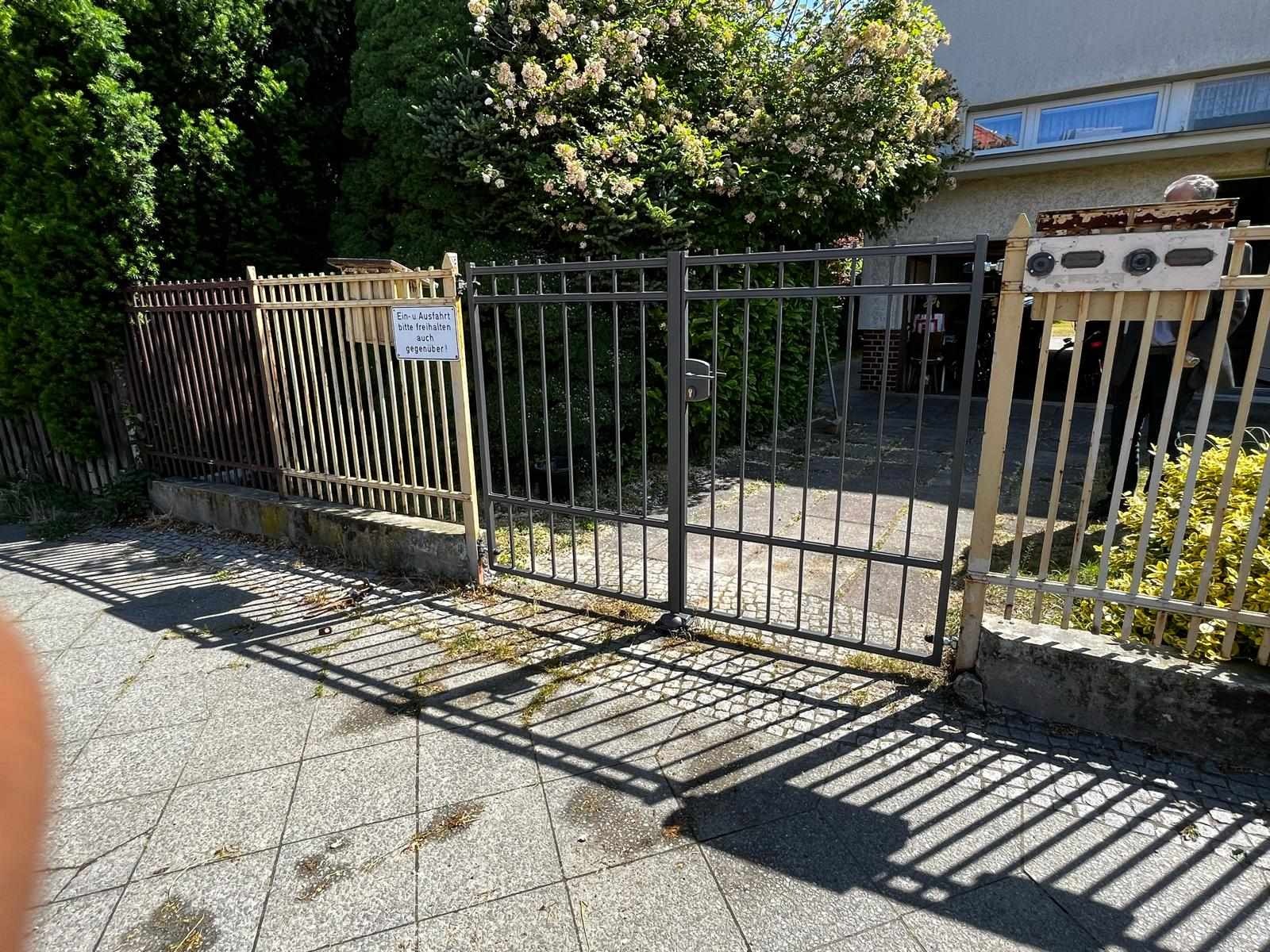 Metalowa brama wjazdowa z tabliczką informacyjną, rzucająca cienie na chodnik z szarej kostki, otoczona zielenią i ogrodzeniami.