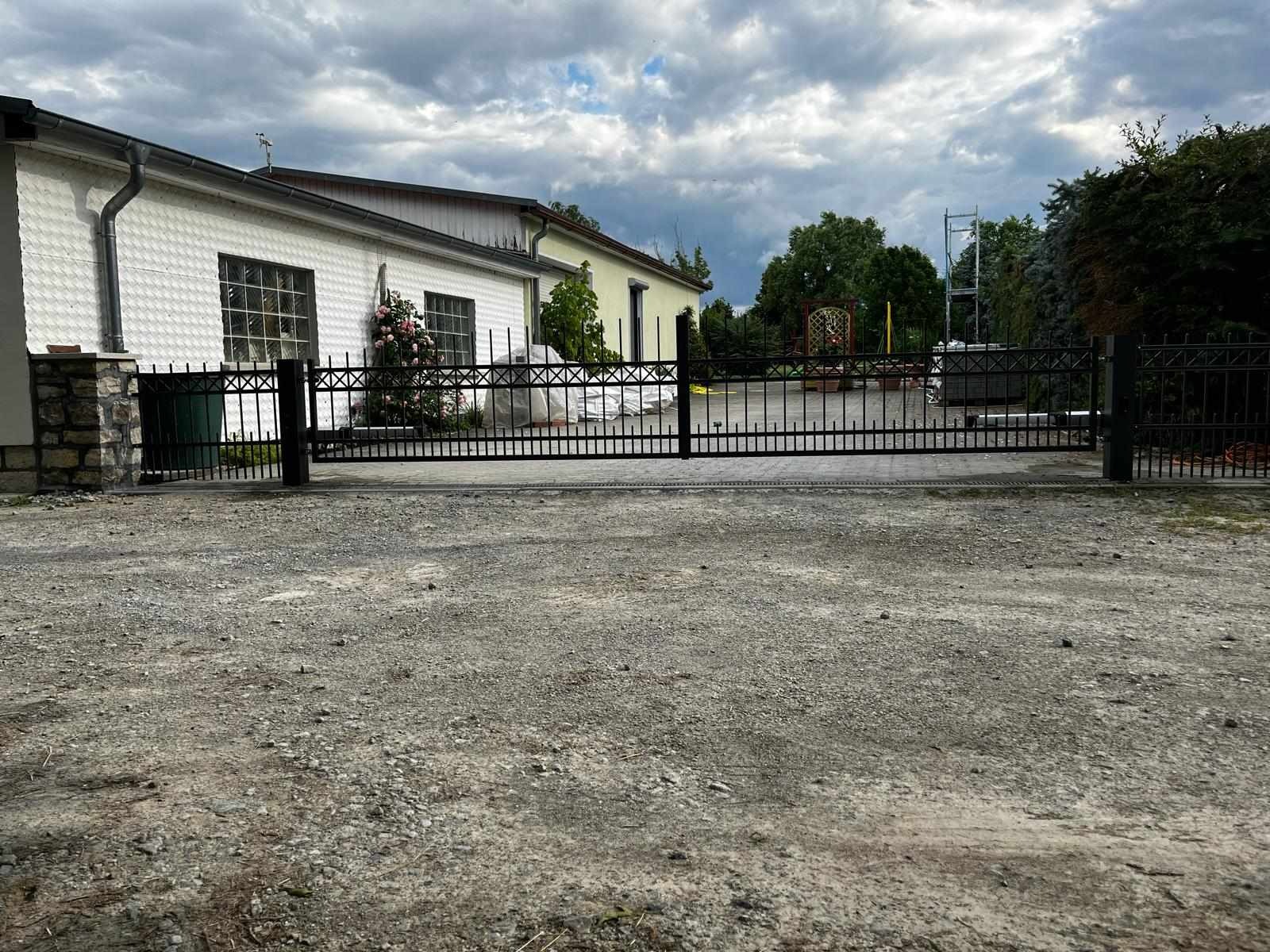 Nowo ułożona kostka brukowa prowadząca do czarnej, metalowej bramy wjazdowej na tle białego budynku z kwitnącymi różami pnącymi się po ścianie.