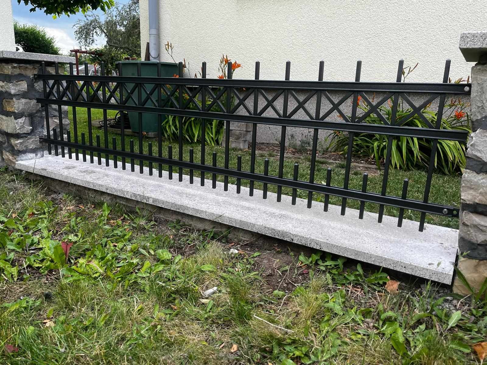 Czarny metalowy płot z dekoracyjnymi krzyżami, osadzony na betonowej podmurówce, z kamiennymi słupkami, otoczony zielenią i kwiatami lilii.