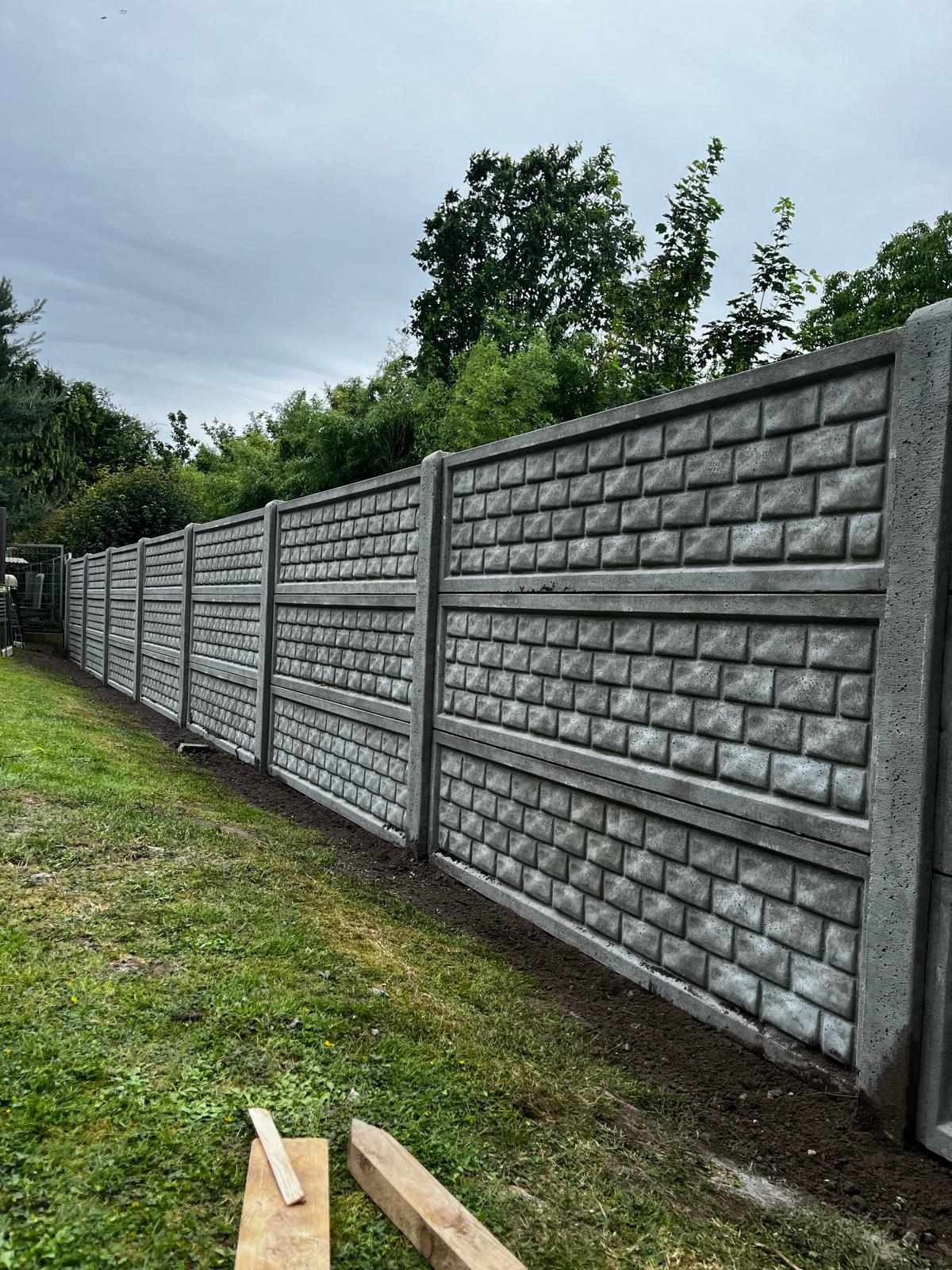 Długi, szary, betonowy płot panelowy z fakturą cegły, oddzielający posesję od zieleni, z drewnianymi elementami leżącymi u podstawy.
