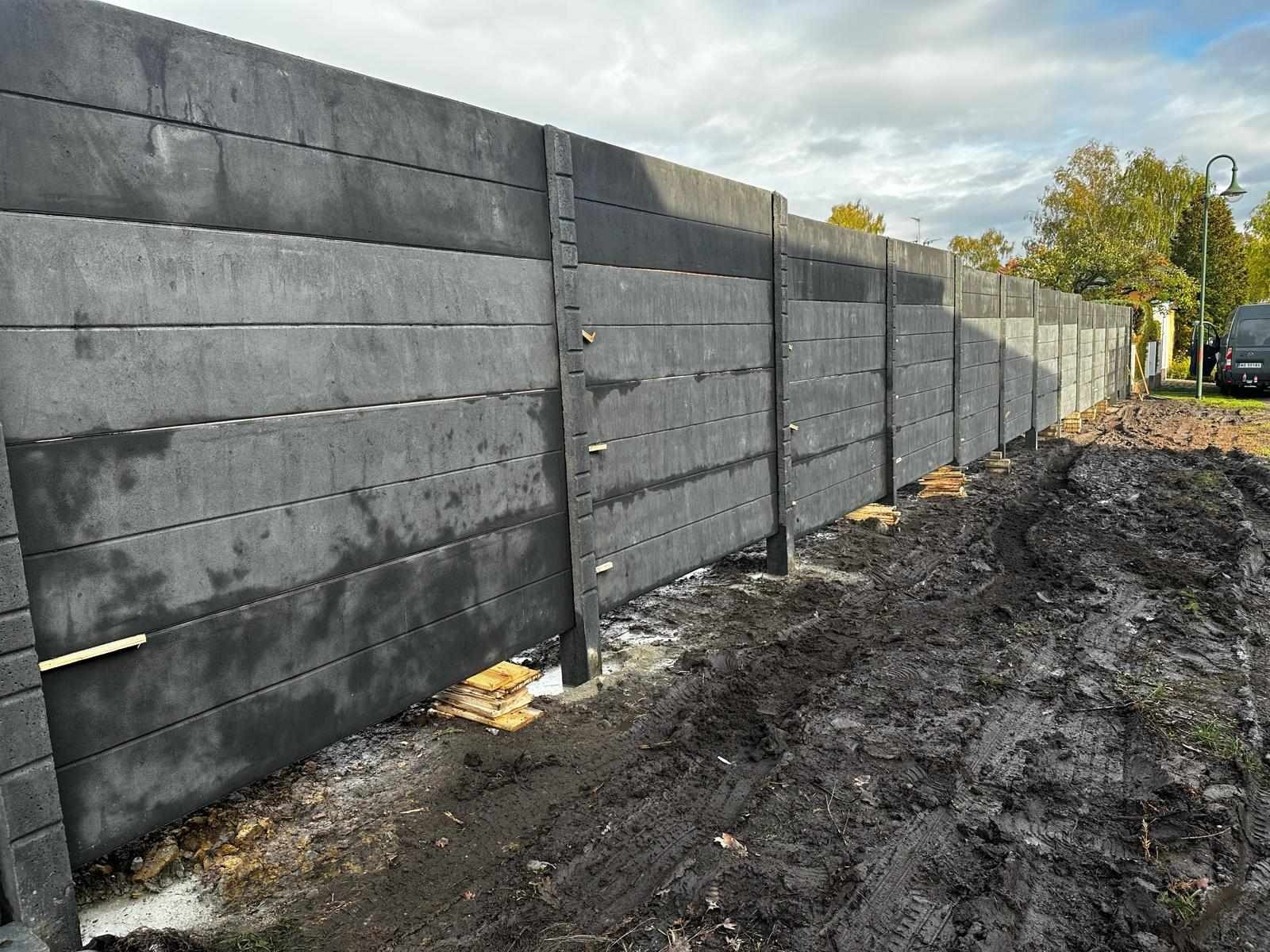 Montaż prefabrykowanego, betonowego ogrodzenia panelowego w trakcie realizacji, widoczne deski stabilizujące i błoto po pracach ziemnych.