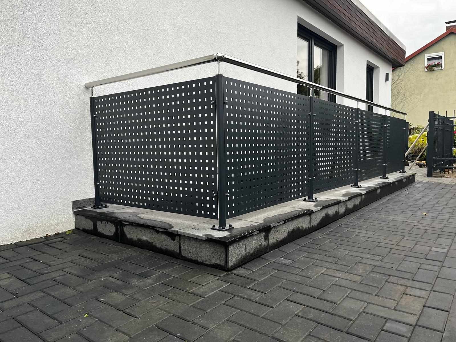 Nowoczesne, ażurowe ogrodzenie panelowe w kolorze grafitowym z metalową poręczą, zamontowane na podmurówce z szarej kostki, otoczone brukowaną nawierzchnią.