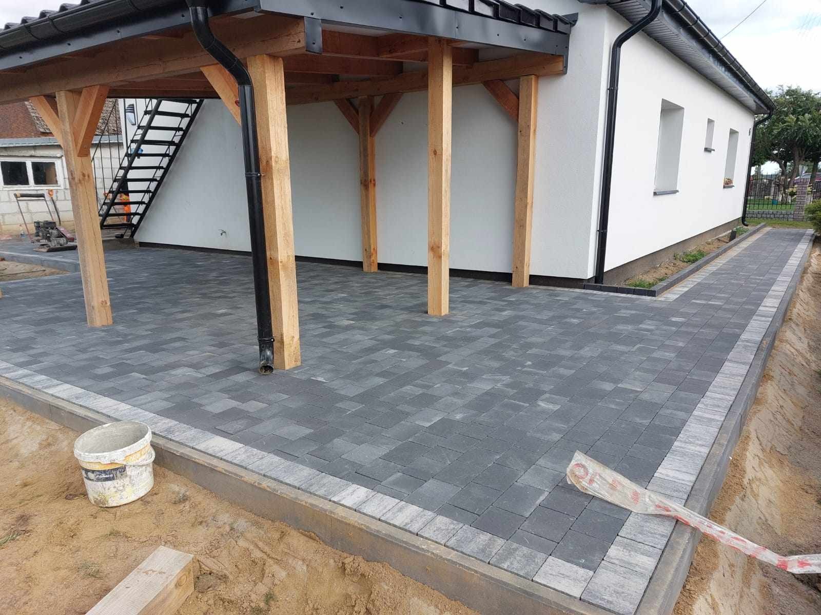 Ułożona szara kostka brukowa przed domem z drewnianą wiatą, widoczny biały plastikowy kubeł i łopata w folii ochronnej.