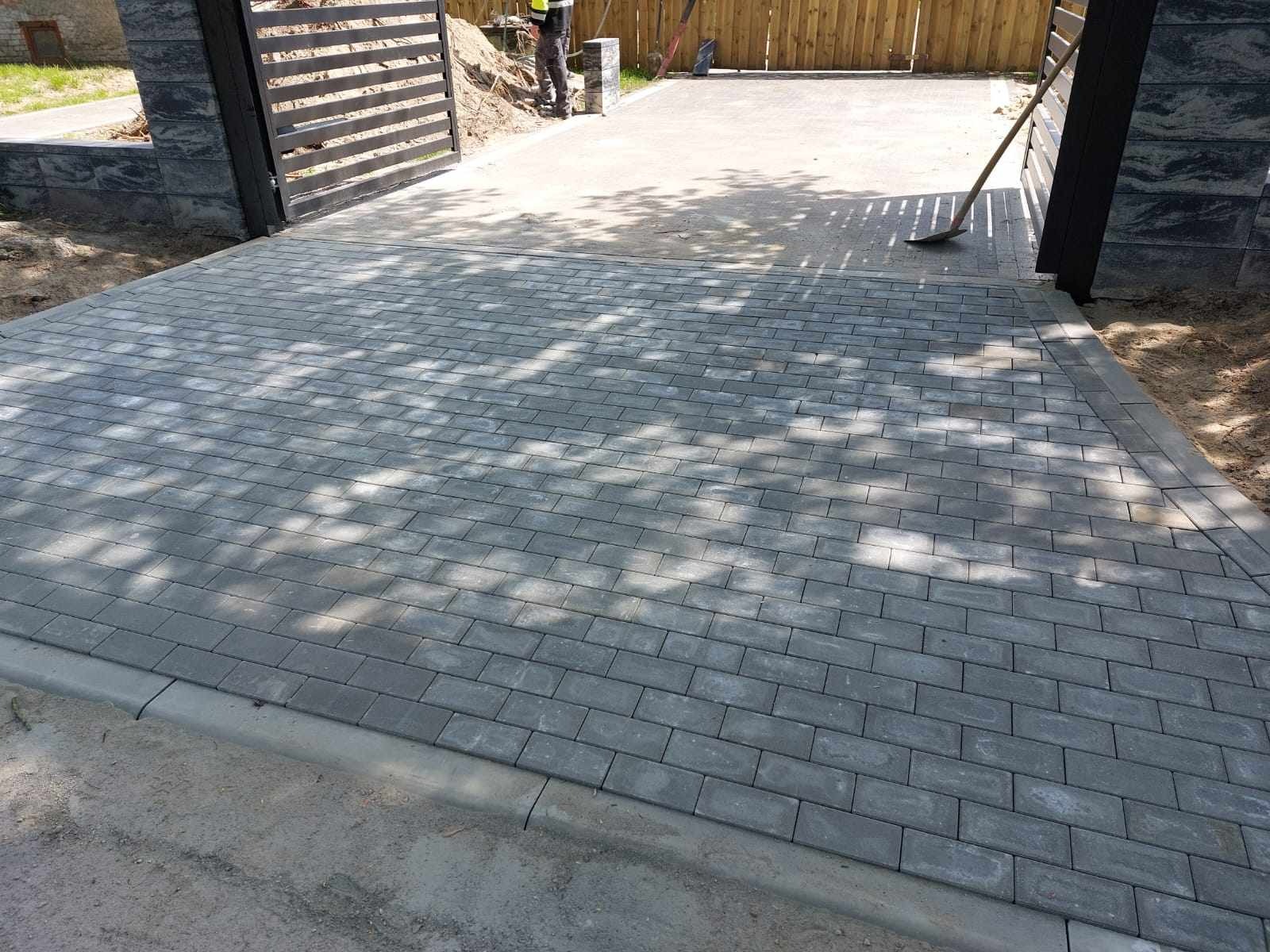 Ułożona szara kostka brukowa w kształcie prostokątów, obrzeża betonowe, brama wjazdowa z czarnymi metalowymi przęsłami, widoczny fragment pracownika z łopatą w tle.
