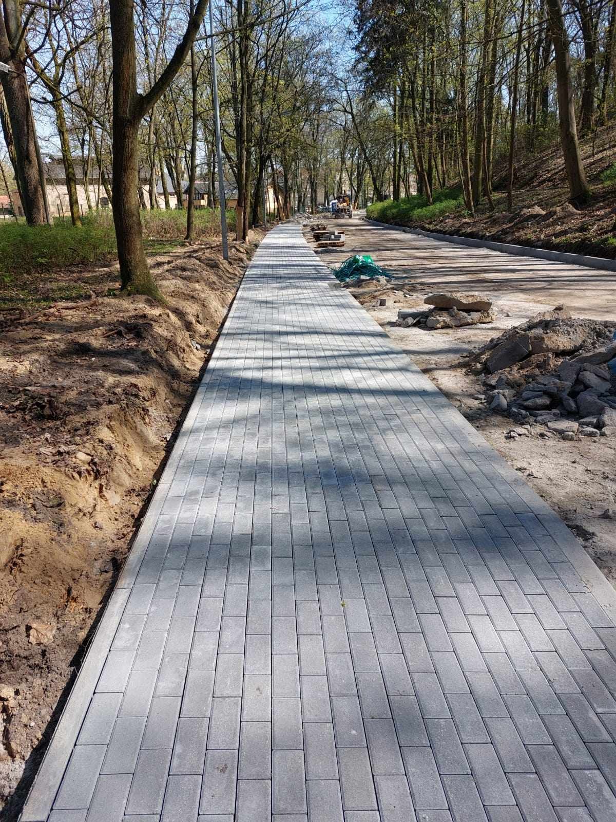 Długa, szara ścieżka z kostki brukowej w trakcie budowy w Kamieniu Wielkim, otoczona drzewami i przygotowanym podłożem.
