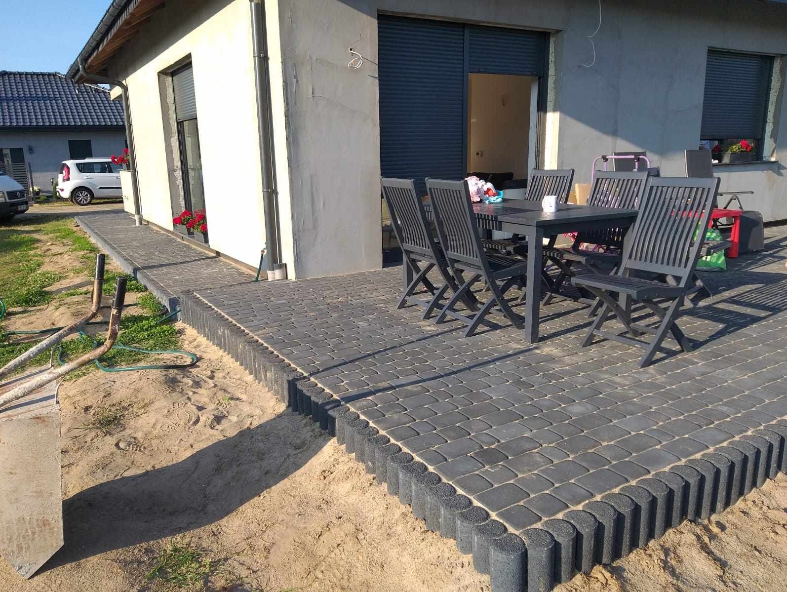 Nowoczesny taras z ciemnej kostki brukowej obramowany palisadą, z kompletem mebli ogrodowych i widokiem na dom.