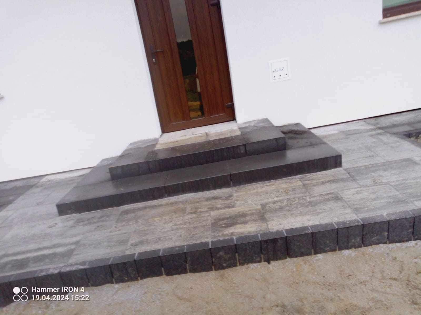 Schody wejściowe do domu wyłożone szarą kostką brukową, z obrzeżem z ciemnej kostki, widok z dołu, obok drzwi wejściowych i skrzynki gazowej.