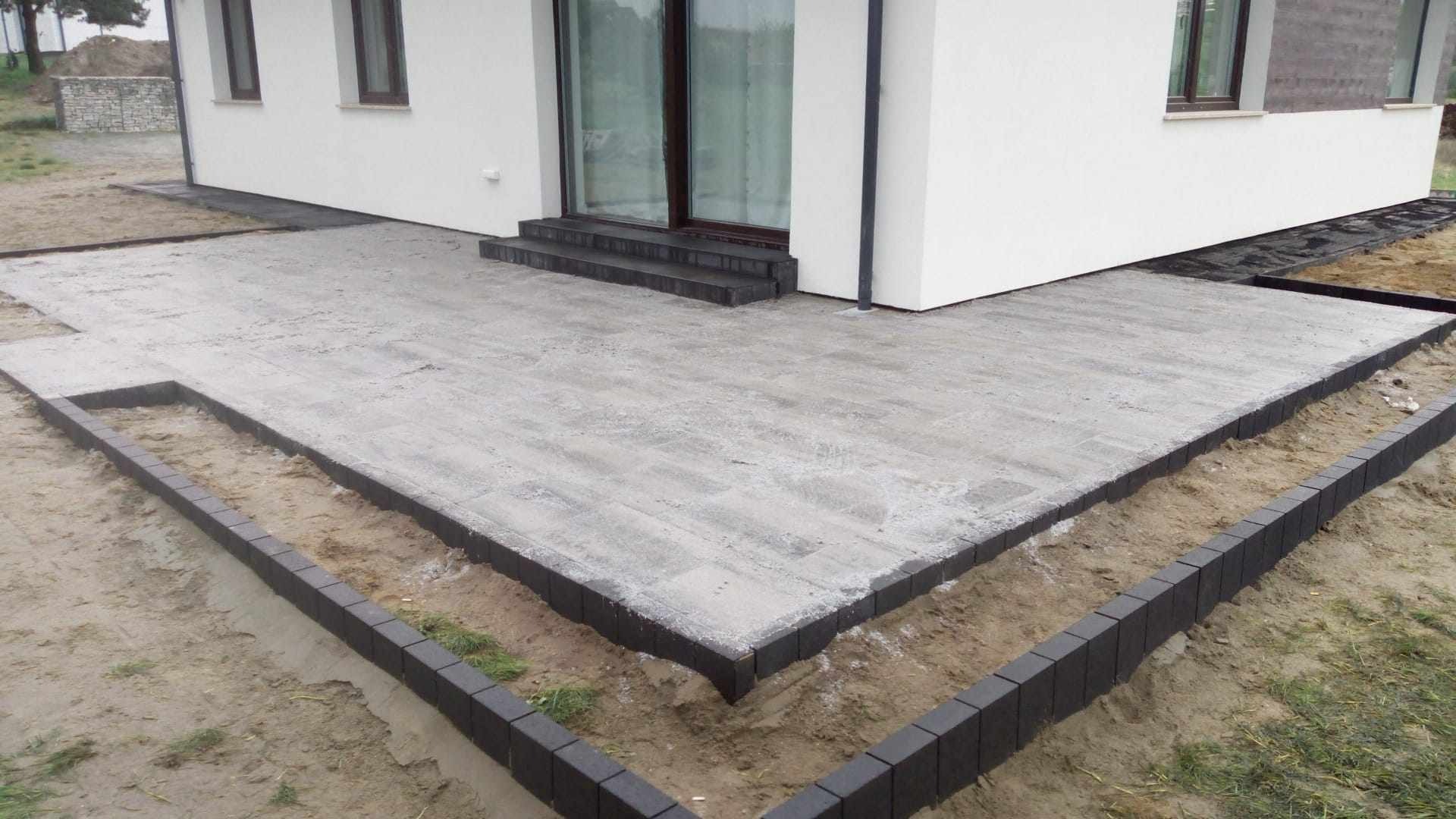Przygotowany podkład pod kostkę brukową z obrzeżami, widoczne czarne obrzeża i szare płyty podkładowe, fragment domu z oknami w tle.
