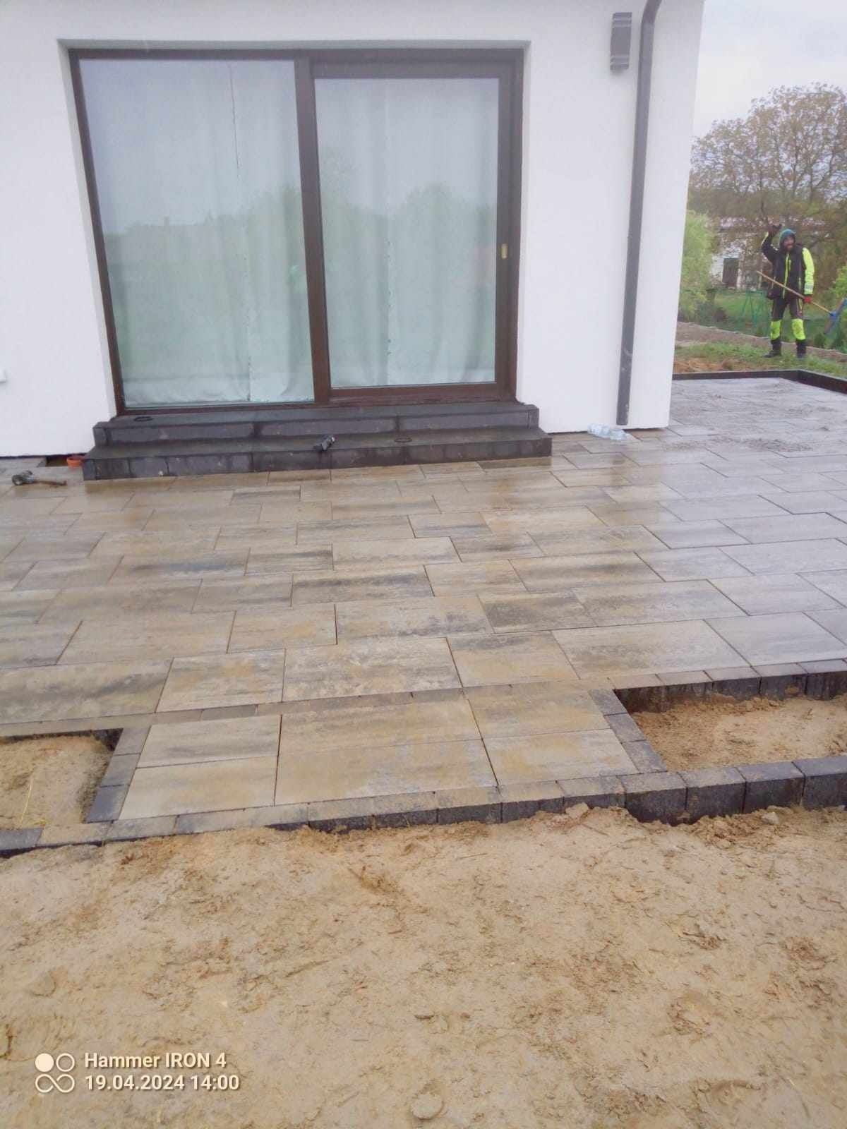 Fragment tarasu z ułożoną częścią szarej kostki brukowej, widoczne przygotowane podłoże z piasku, w tle biała ściana budynku z dużym oknem przesuwnym i pracownik z grabiami.