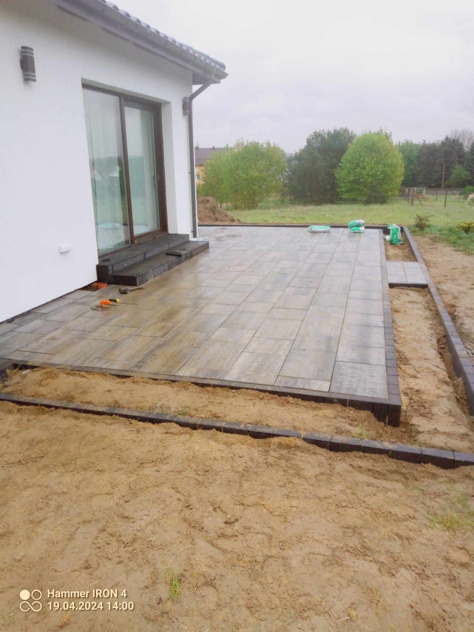 Układanie szarych płyt tarasowych z ciemną obwódką przy domu, widoczne narzędzia i worki z materiałem budowlanym.