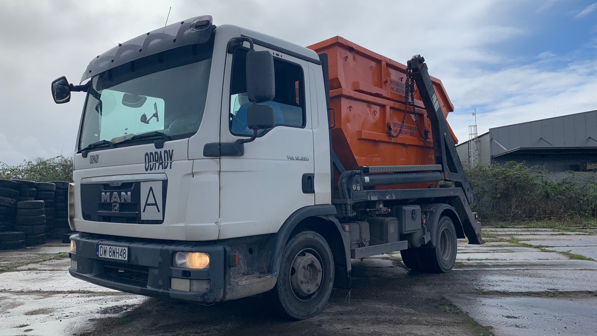 Ciężarówka MAN z pomarańczowym kontenerem na platformie, zaparkowana na betonowym placu z widocznymi stertami opon w tle.