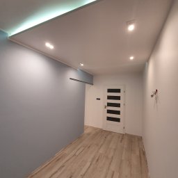 Dawid bonk - Wnętrze korytarza po remoncie: ściana pomalowana na szaro, biała ściana z zamontowanymi gniazdkami, jasna podłoga imitująca drewno, białe drzwi z czarnymi wstawkami i oświetlenie sufitowe LED.