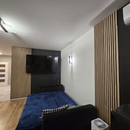 Dawid bonk - Nowoczesny pokój z drewnianymi lamelami na ścianach, czarną ścianą za telewizorem i klimatyzatorem, kanapą z niebieską narzutą, widoczny fragment podłogi z panelami oraz fragment korytarza...