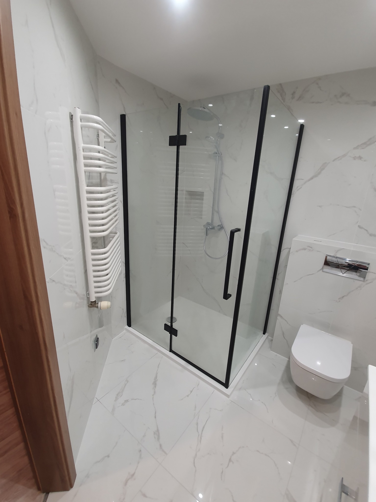 Nowoczesna łazienka z kabiną prysznicową w czarnej ramie, marmurowe płytki na ścianach i podłodze, wisząca toaleta i grzejnik drabinkowy.