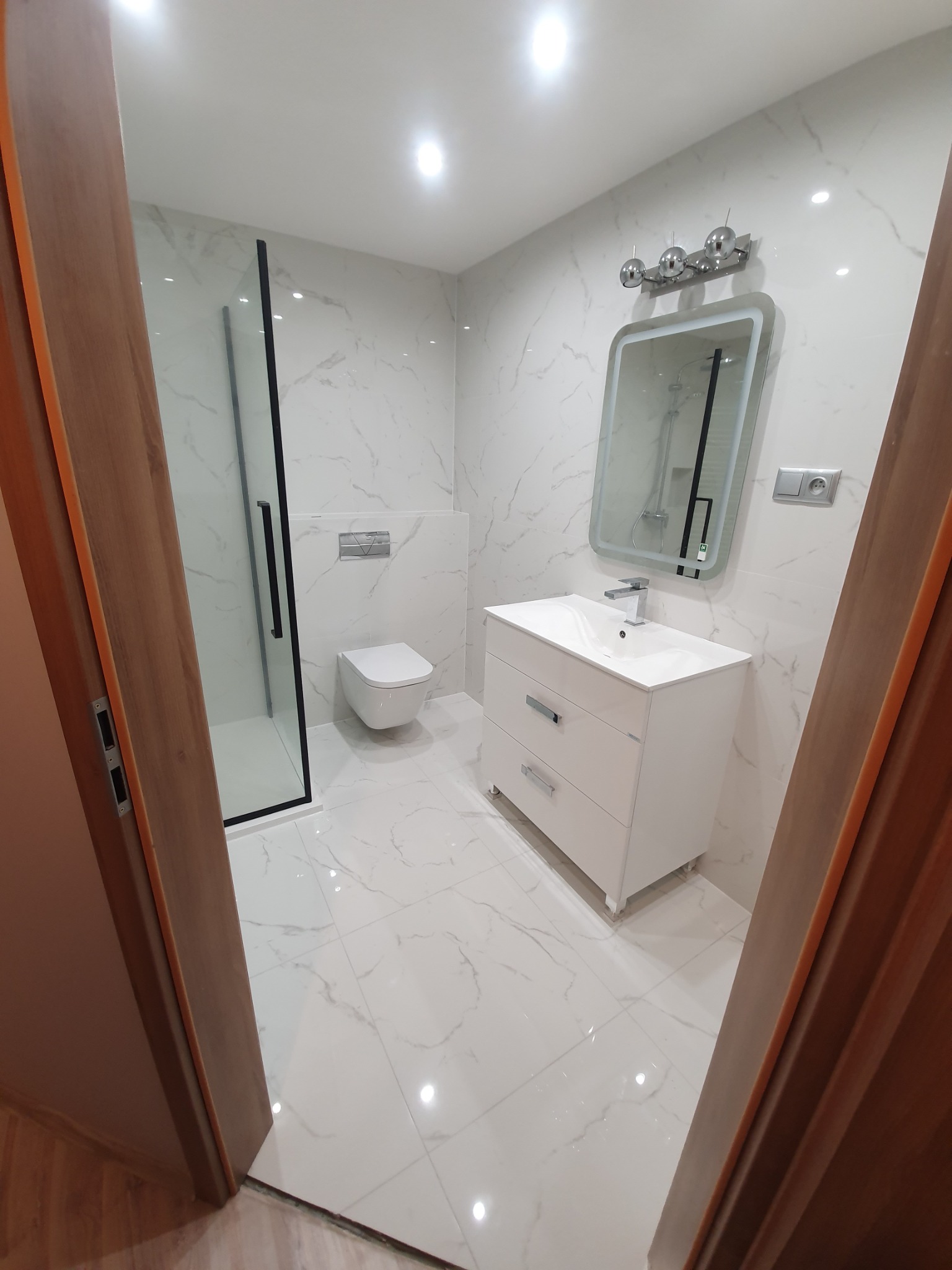 Nowoczesna łazienka z białymi marmurkowymi płytkami, kabiną prysznicową z czarnymi ramami, podwieszaną toaletą i białą szafką umywalkową z trzema szufladami, widziana zza drewnianej ościeżnicy drzwi.