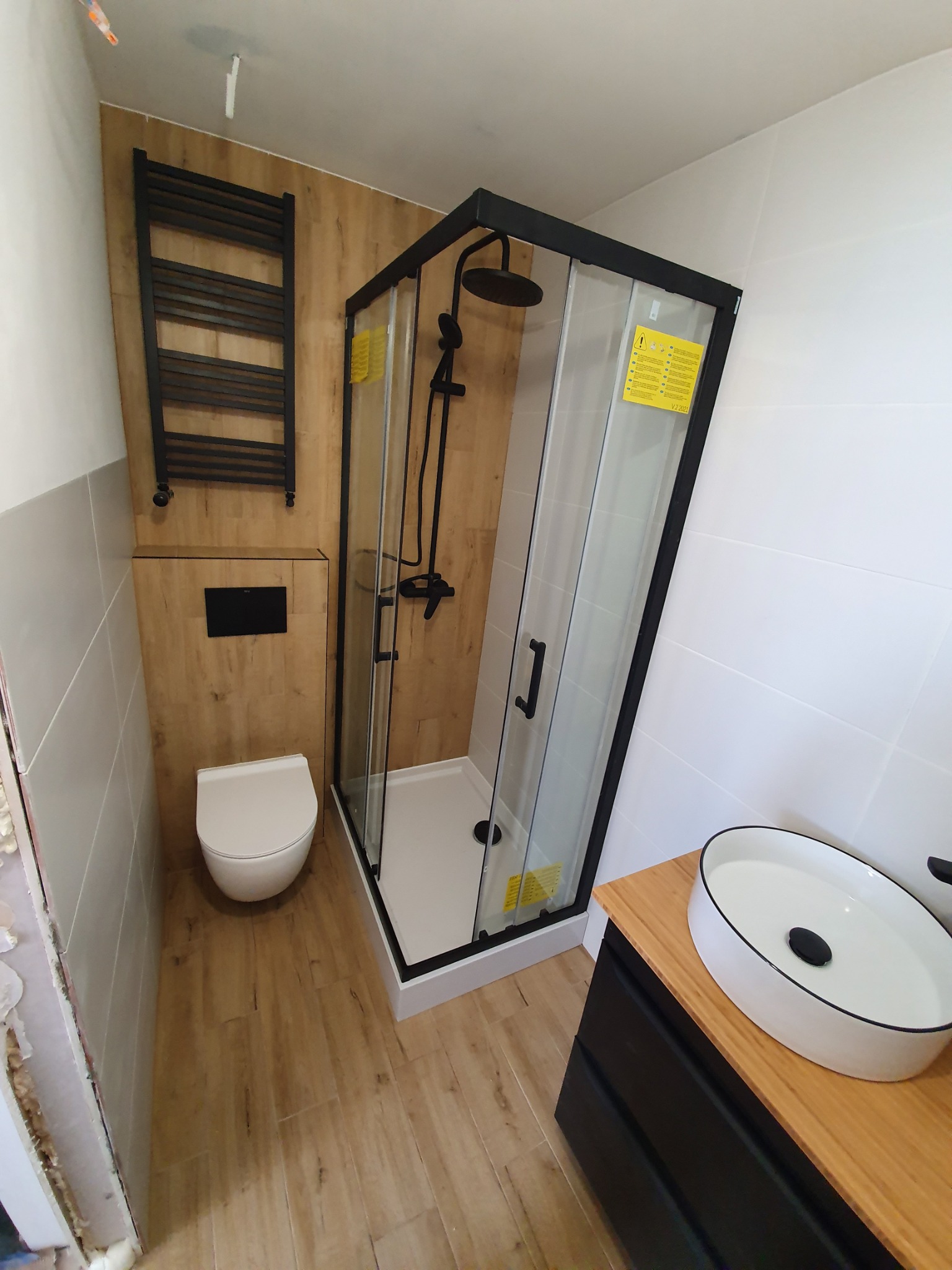 Nowoczesna łazienka z kabiną prysznicową w czarnej ramie, wiszącą toaletą, grzejnikiem drabinkowym i umywalką nablatową, wykończona płytkami imitującymi drewno i białymi płytkami.