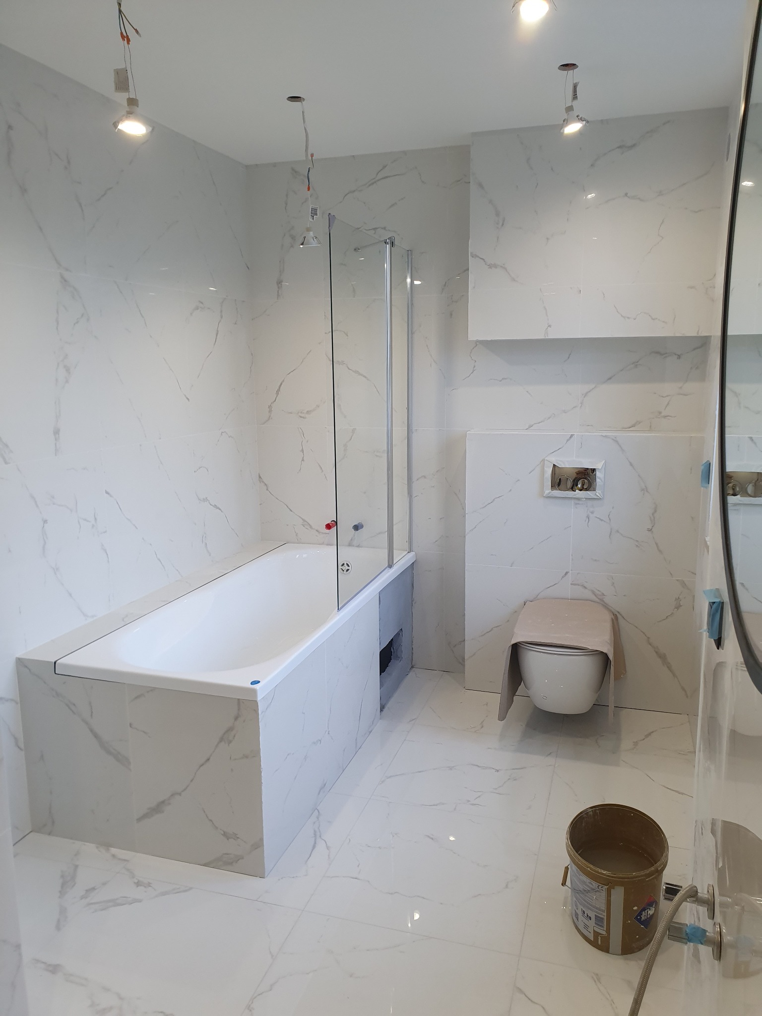 Biała łazienka w trakcie remontu z wanną z parawanem, toaletą przykrytą materiałem ochronnym, widoczne niedokończone oświetlenie i wiadro z zaprawą na podłodze.