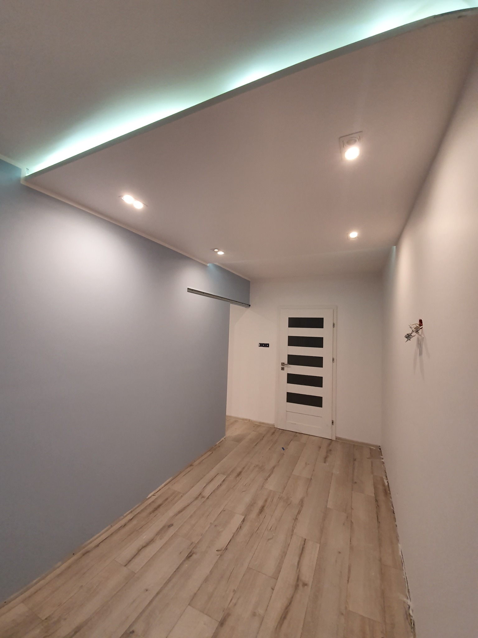 Wnętrze korytarza po remoncie: ściana pomalowana na szaro, biała ściana z zamontowanymi gniazdkami, jasna podłoga imitująca drewno, białe drzwi z czarnymi wstawkami i oświetlenie sufitowe LED.