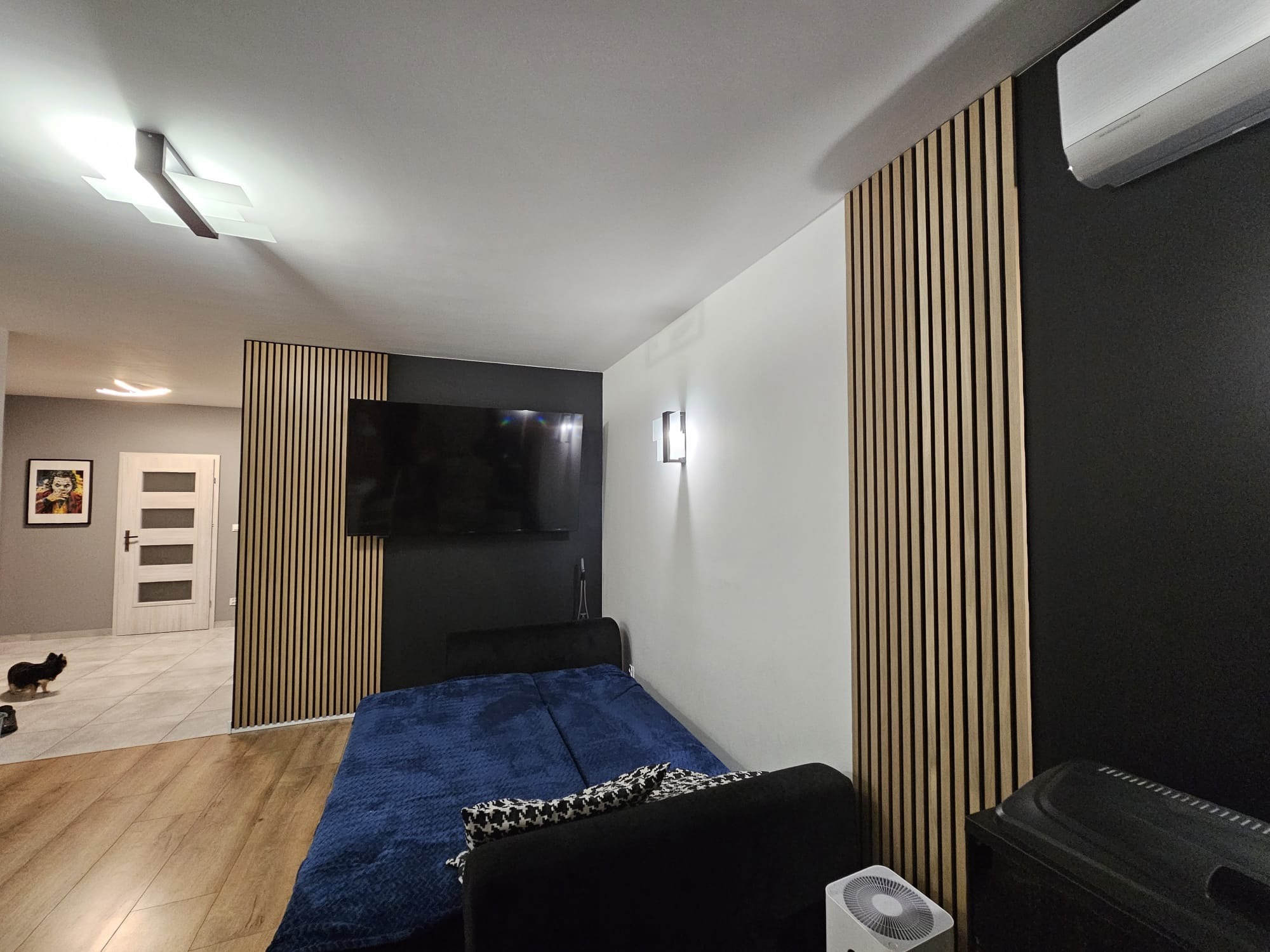 Nowoczesny pokój z drewnianymi lamelami na ścianach, czarną ścianą za telewizorem i klimatyzatorem, kanapą z niebieską narzutą, widoczny fragment podłogi z panelami oraz fragment korytarza...