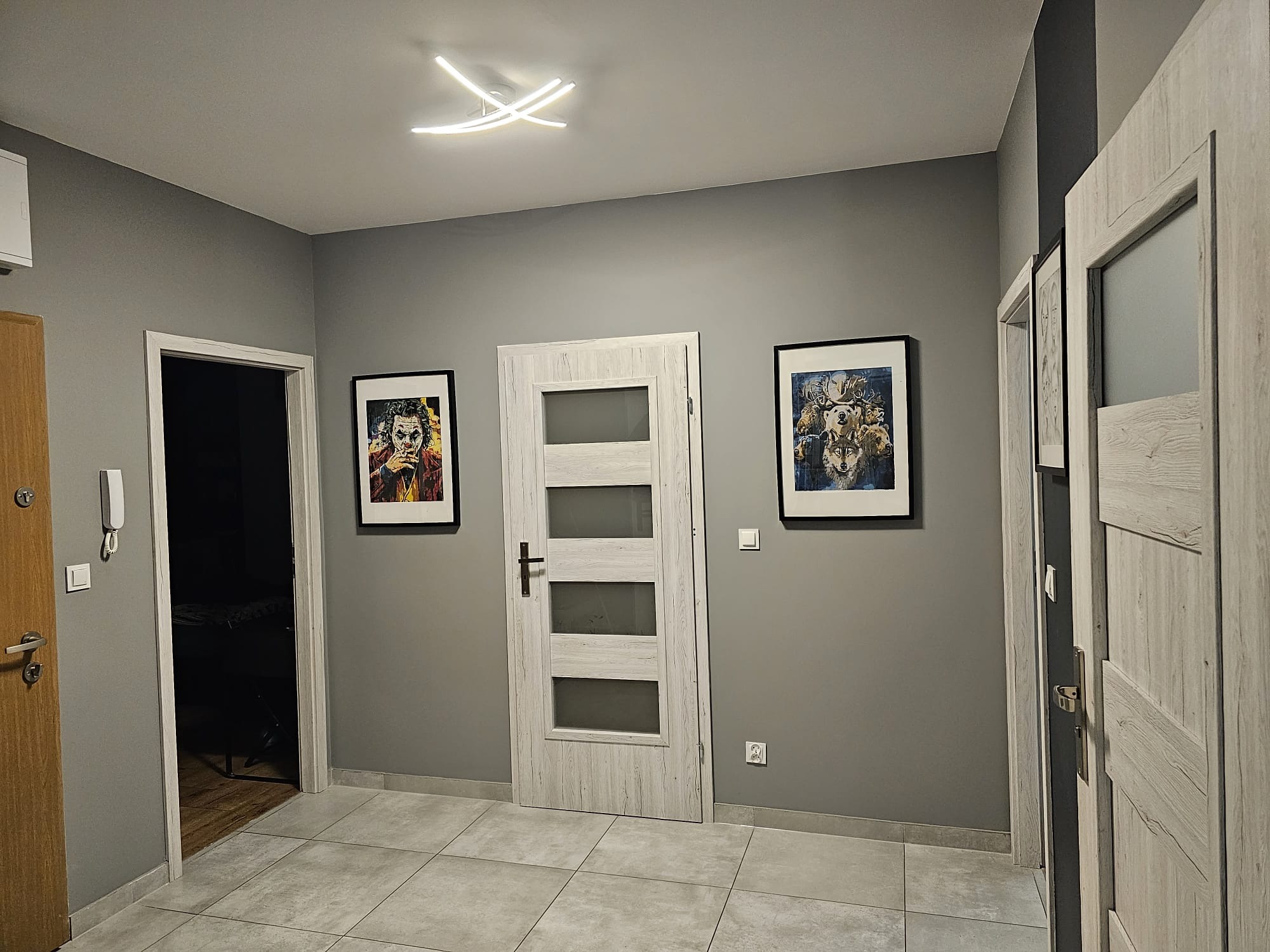 Szare wnętrze przedpokoju z trzema parami drzwi, podłogą z szarych płytek, oświetleniem sufitowym LED, dwoma obrazami w czarnych ramach i szarą ścianą.