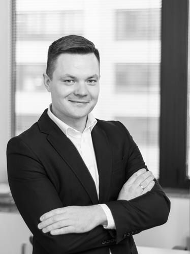 Portret biznesowy mężczyzny w garniturze, z założonymi rękami, uśmiechniętego, na tle okna z żaluzjami, w czerni i bieli.